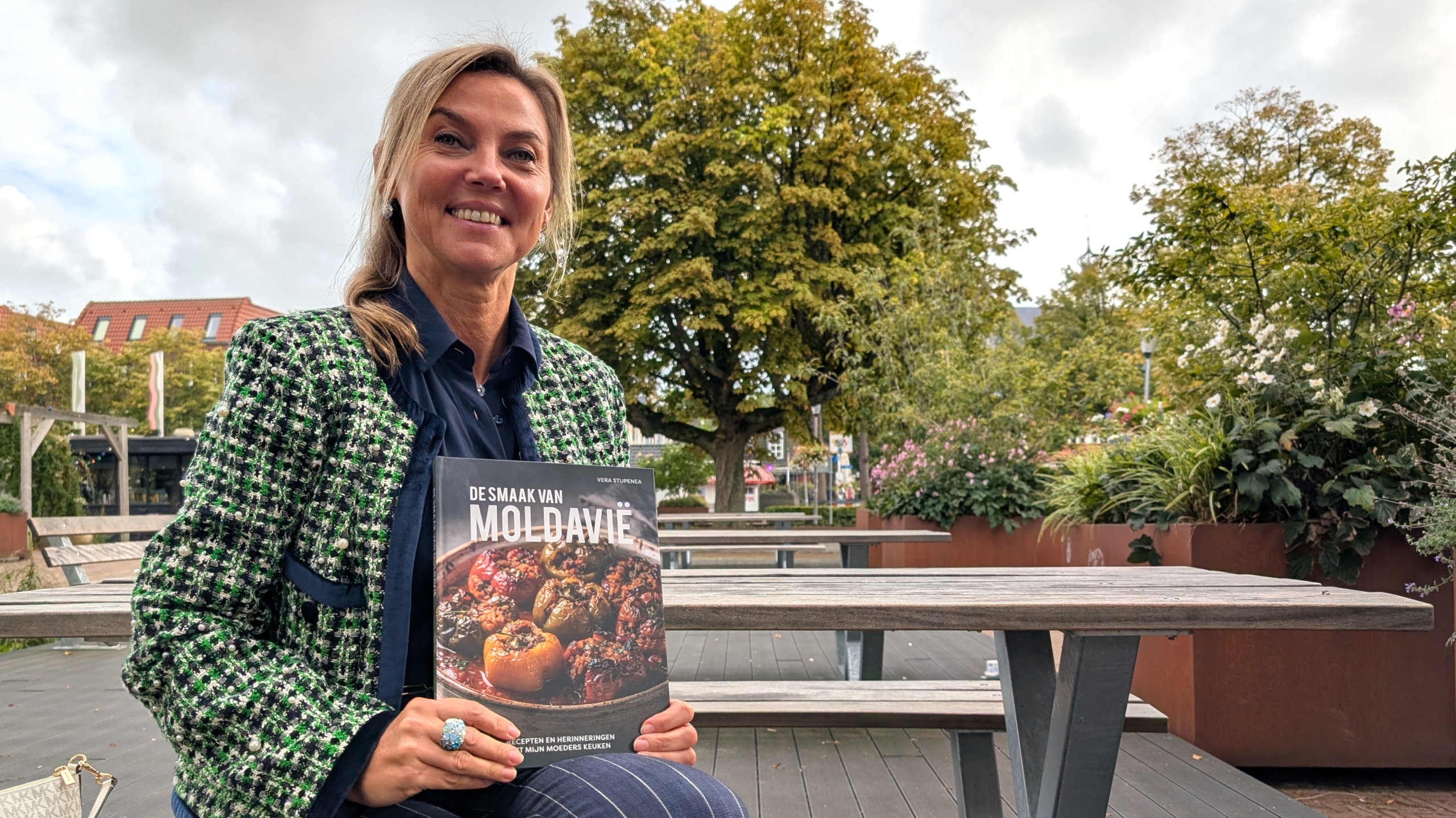 Vrouw in een park houdt het kookboek De smaak van Moldavië vast, zittend bij een picknicktafel met bomen op de achtergrond