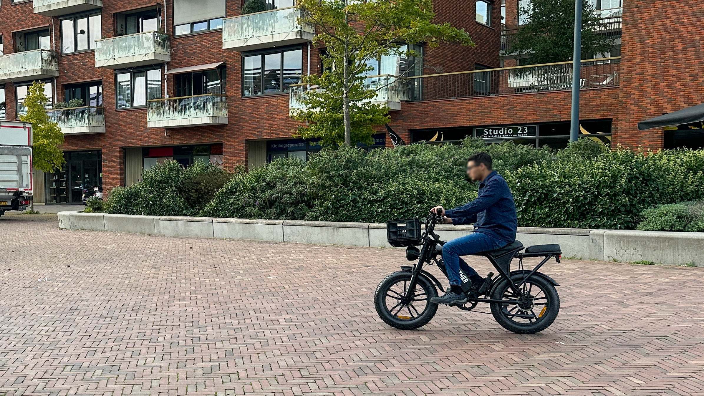 Man rijdt op een zwarte elektrische fiets met mandje langs een bakstenen appartementencomplex met balkons