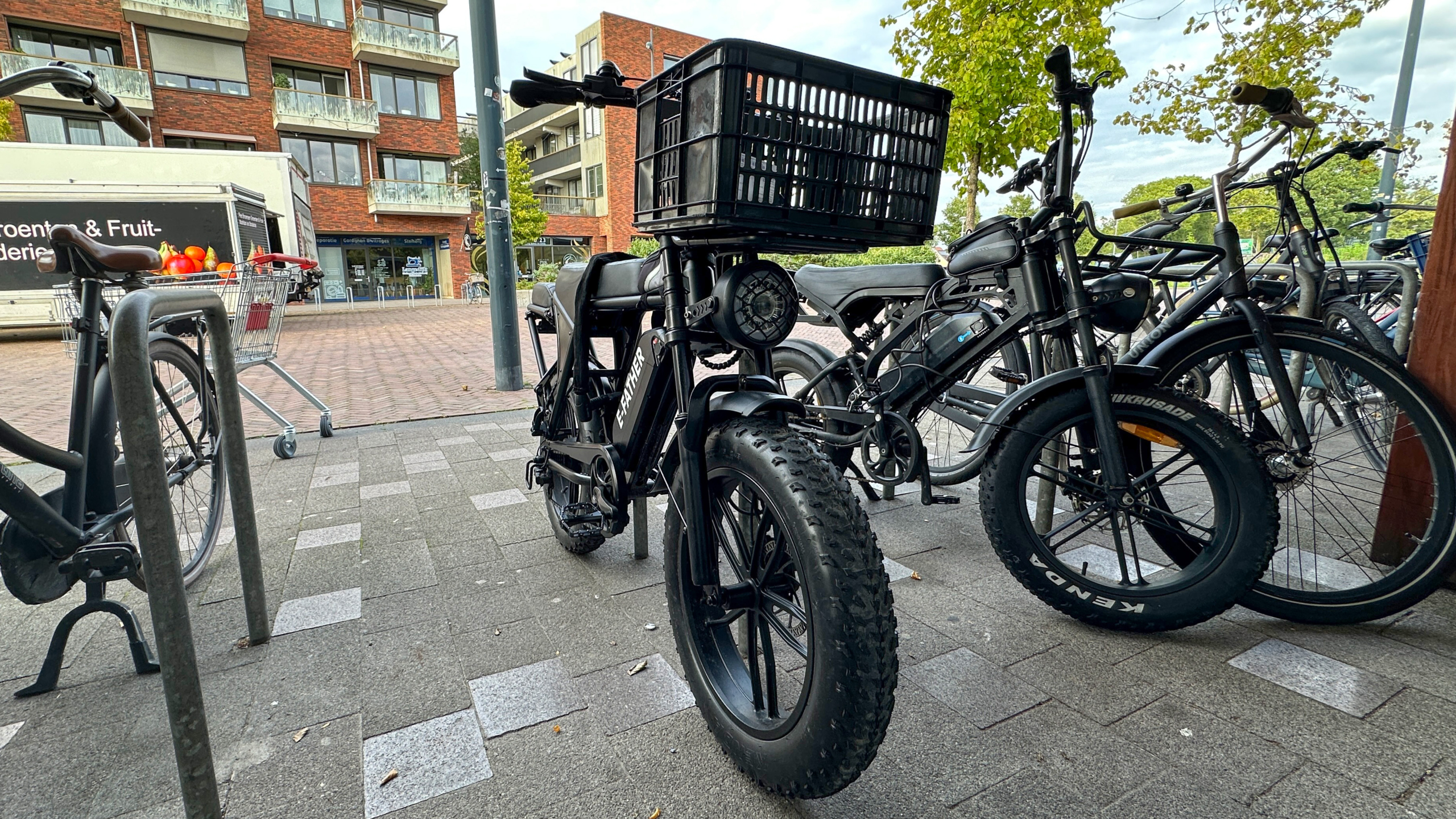 Zwarte e-fatbike met grote mand voorop geparkeerd bij fietsenrekken op een stoep, met flats en bomen op de achtergrond