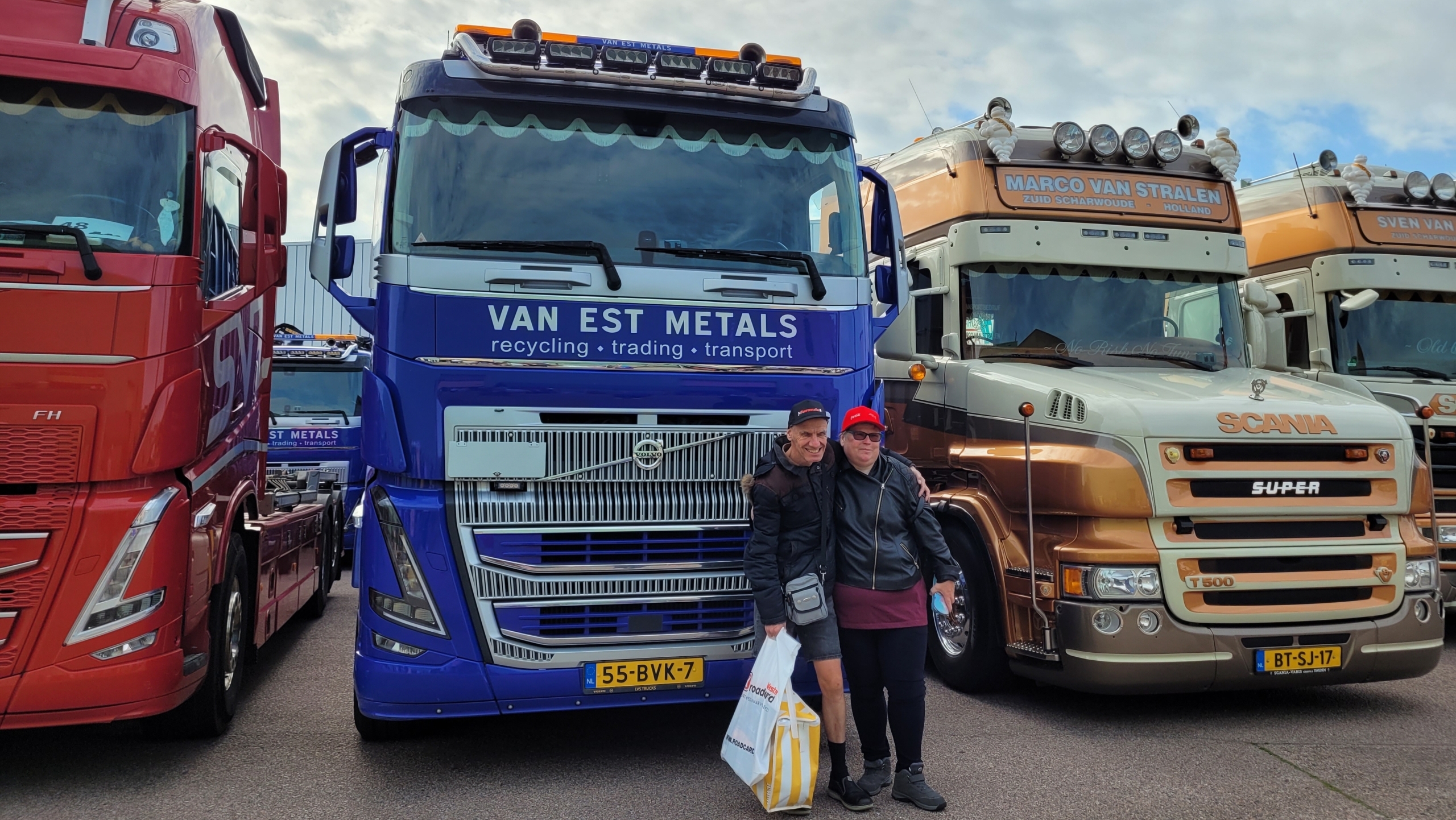 Twee personen poseren voor drie geparkeerde vrachtwagens, met centraal een blauwe truck met de tekst Van Est Metals