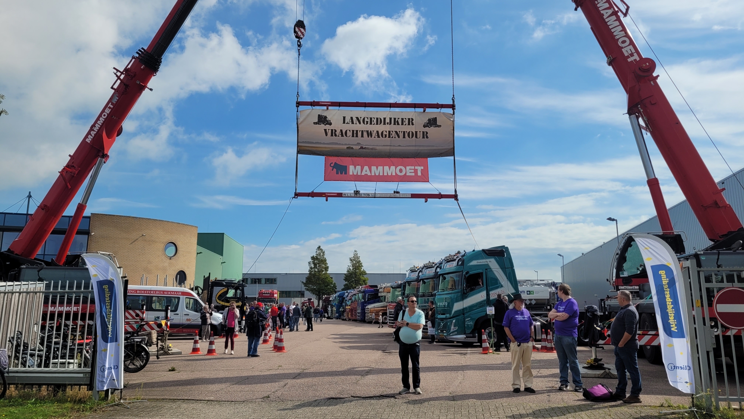 Twee rode Mammoet-kranen hijsen een spandoek “Langedijker Vrachtwagentour” boven een rij vrachtwagens en bezoekers