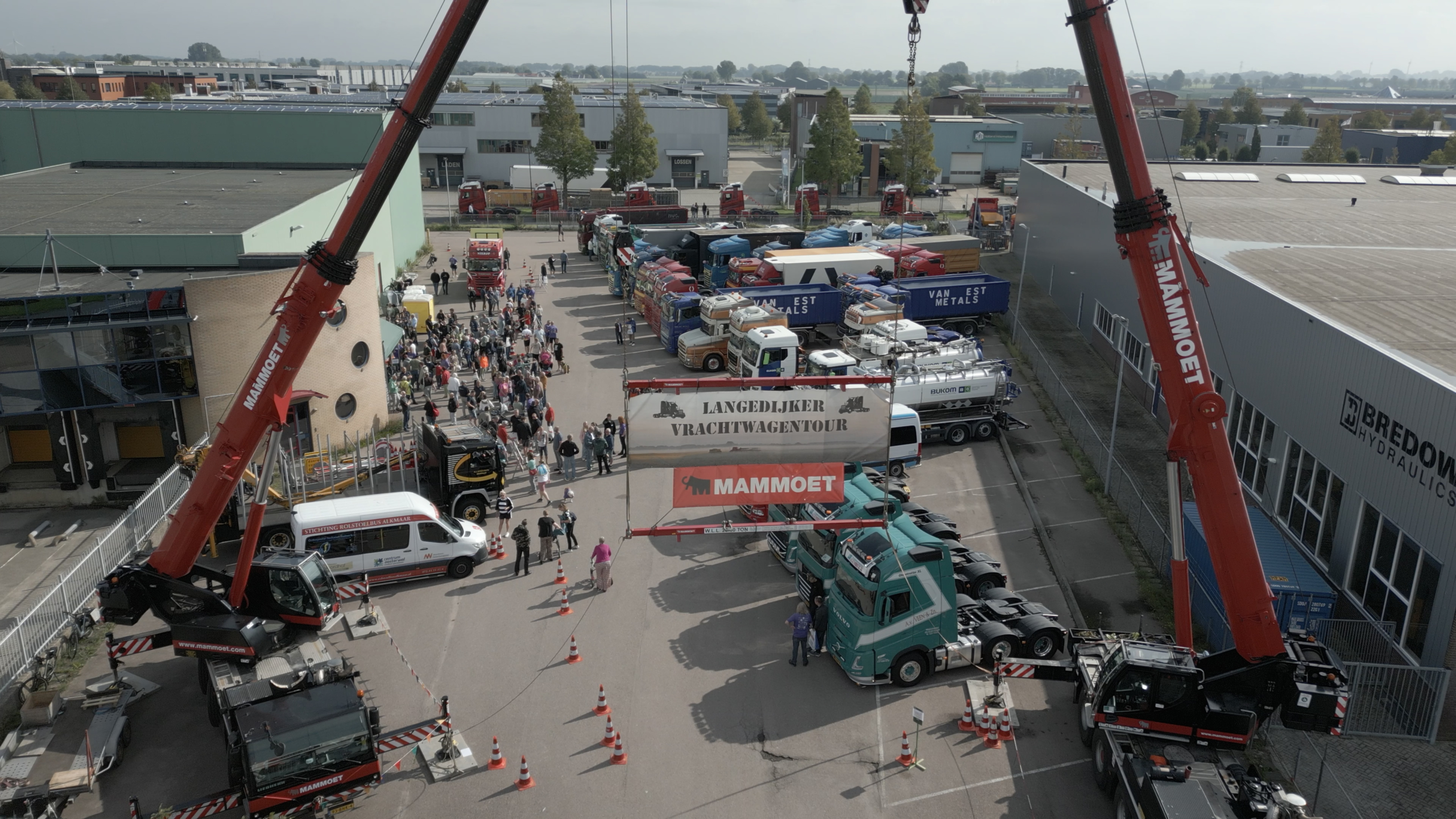 Twee Mammoet-kranen hijsen een spandoek “Langedijker Vrachtwagentour” boven een terrein vol trucks en bezoekers