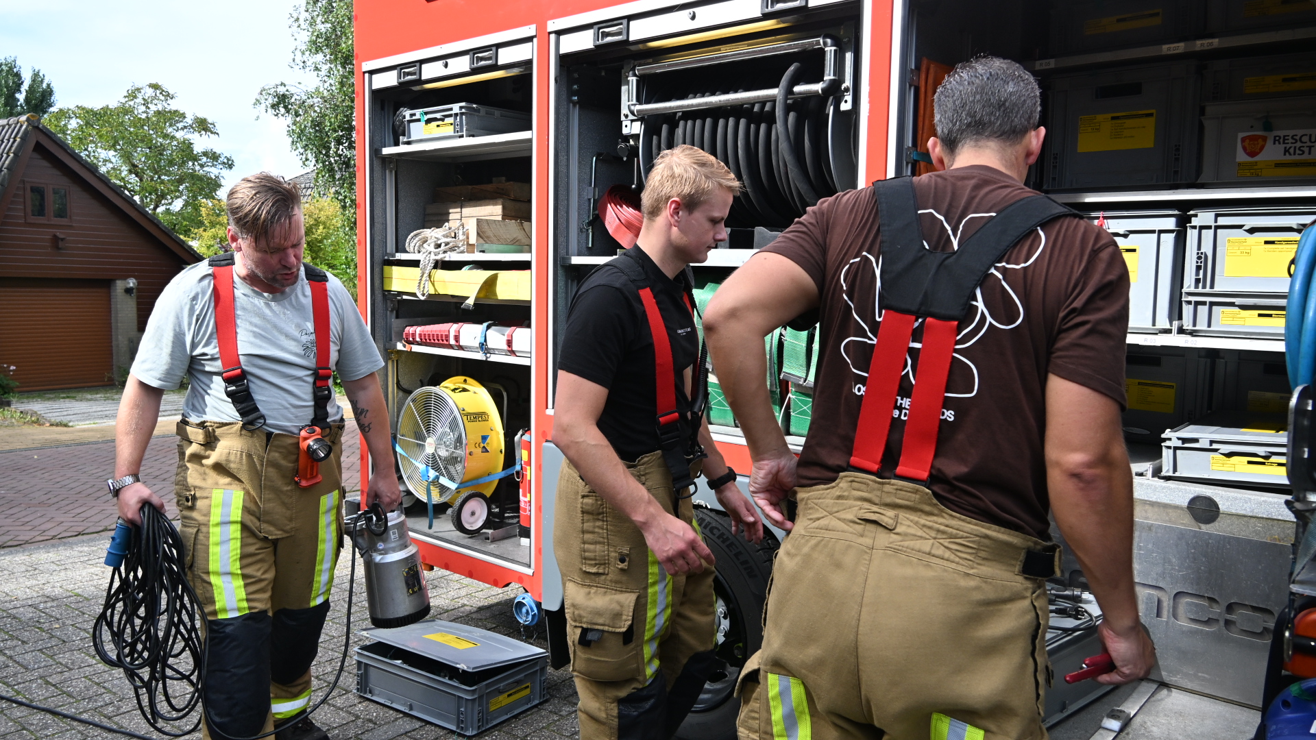 Drie brandweerlieden halen materiaal uit een open brandweerwagen, met slangen, kisten en een ventilator zichtbaar