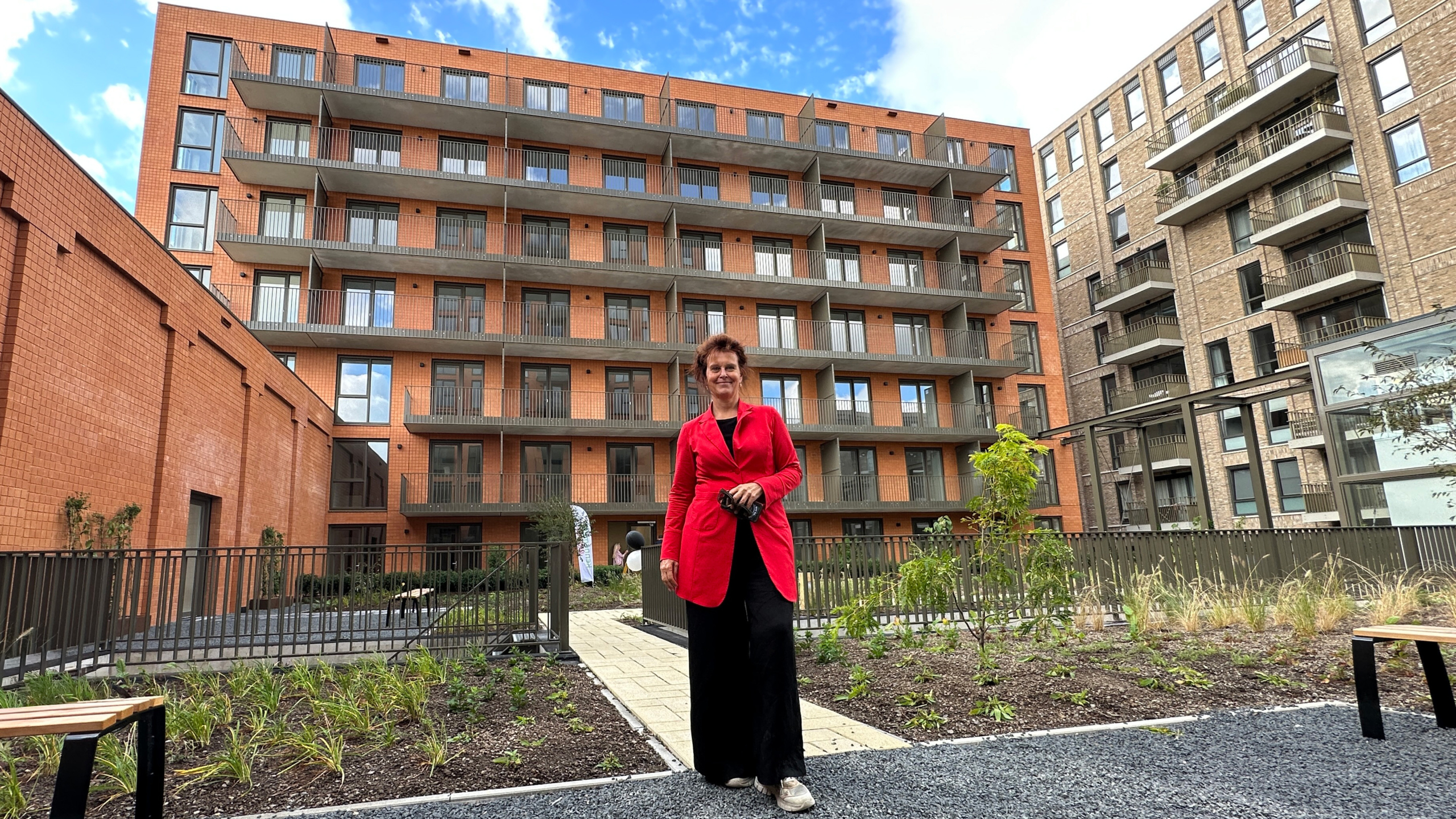Vrouw in rode jas in binnentuin met jonge beplanting, met op de achtergrond een appartementencomplex met balkons