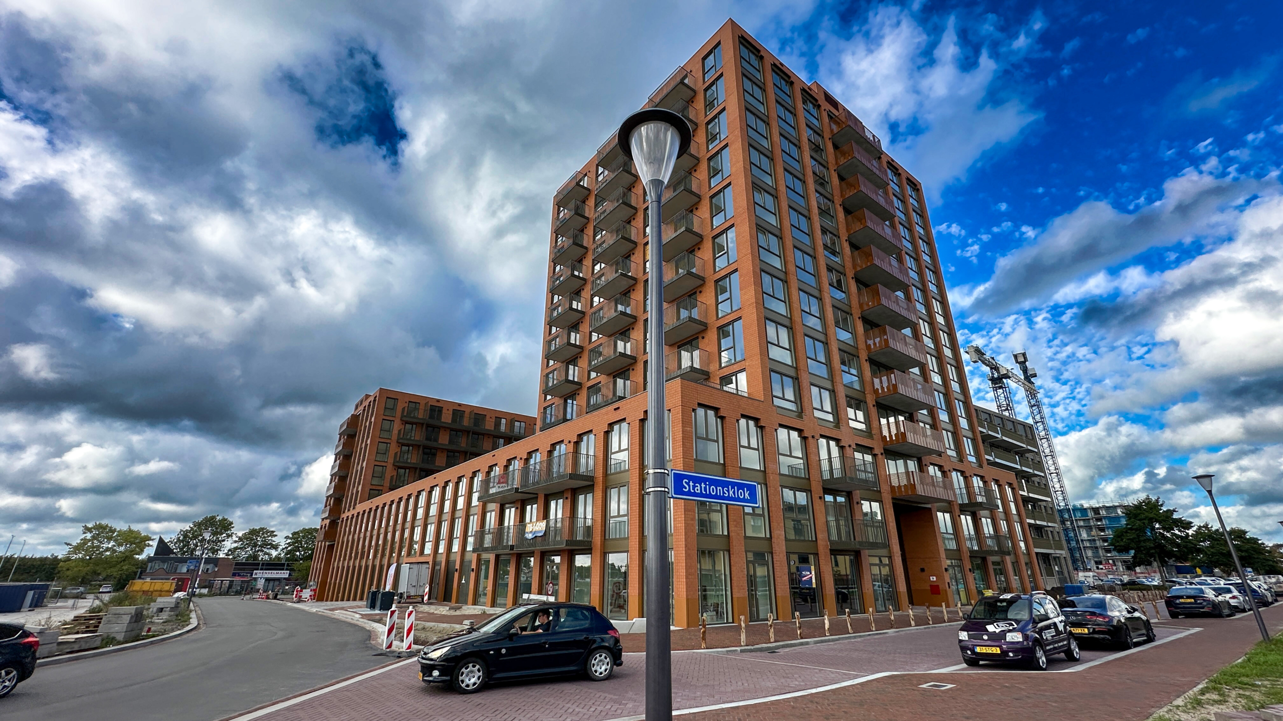 Hoog bakstenen appartementencomplex met balkons aan een kruispunt, straatnaambord ‘Stationsklok’ en wolkenlucht