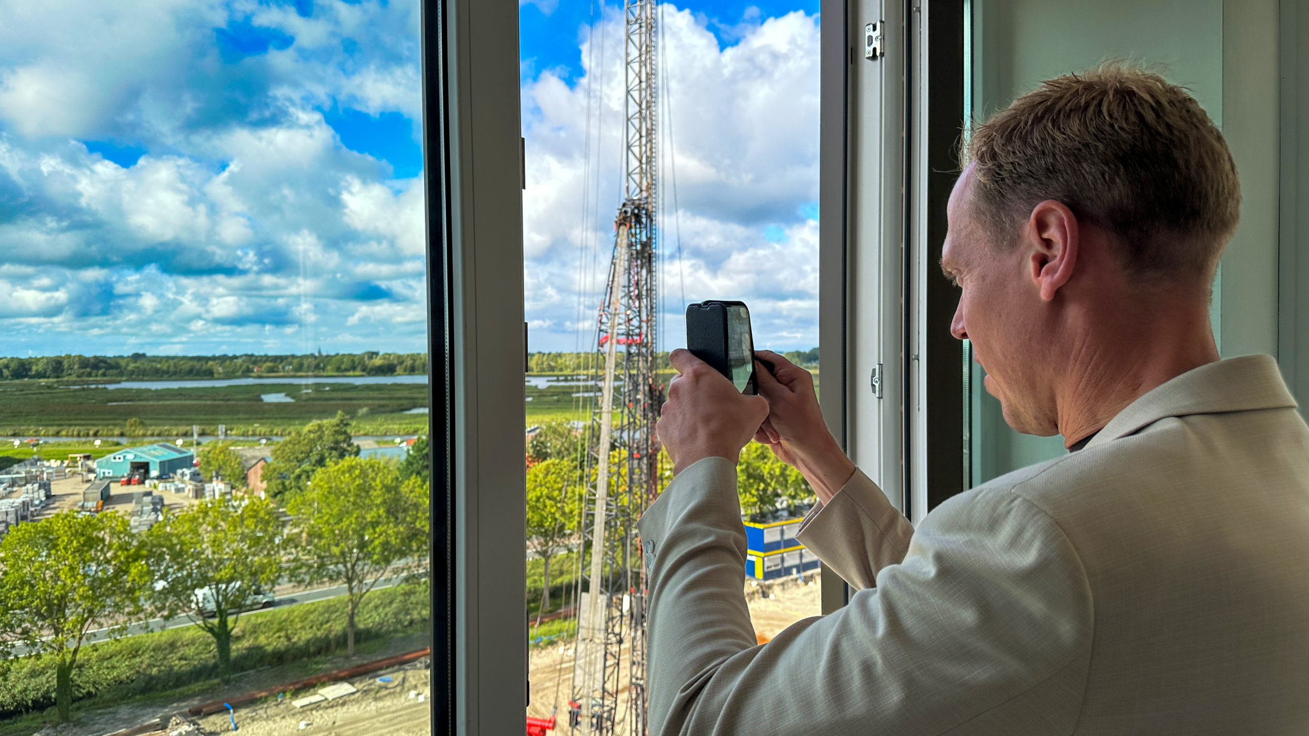 Man bij een raam maakt met zijn smartphone een foto van een hoge bouwkraan en het landschap daarachter