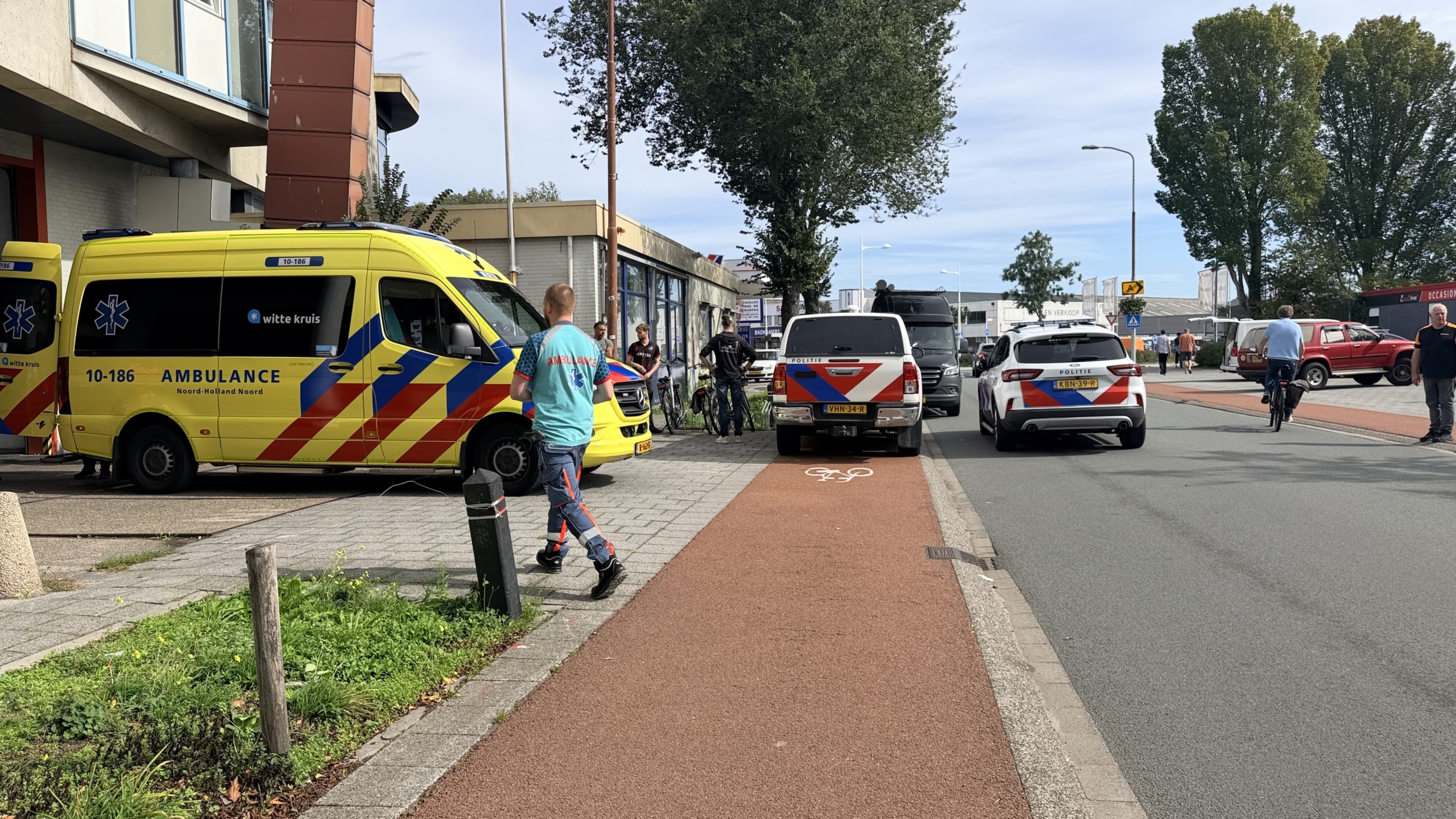 Ambulance en politieauto’s staan langs de weg bij een gebouw, met hulpverlener en omstanders op de stoep