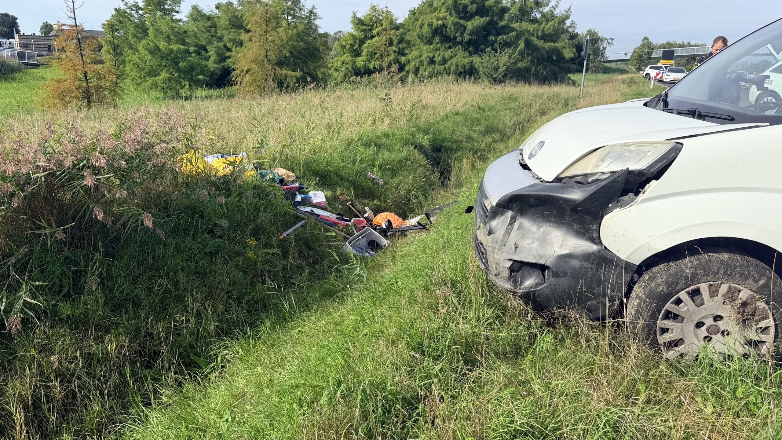 Beschadigde witte bestelwagen in de berm, met verspreide spullen in het gras en een politieauto op de achtergrond