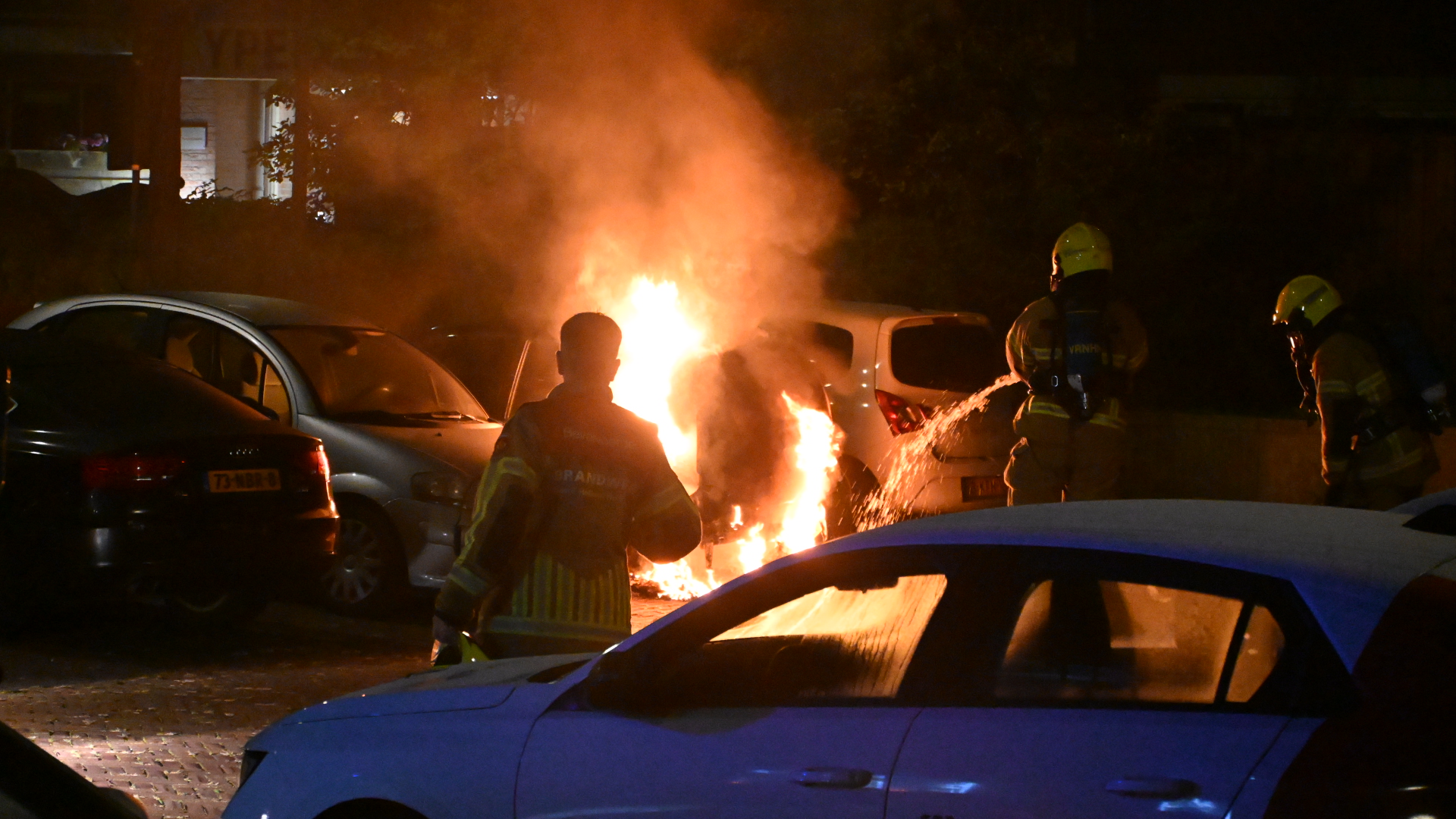 Brandweer blust een brandende auto tussen geparkeerde voertuigen in het donker, met grote vlammen en rook