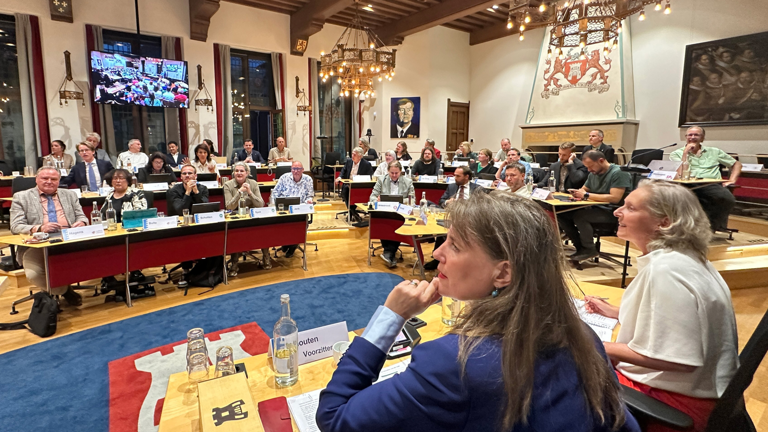 Raadszaal met gemeenteraadsleden aan tafels tijdens vergadering, met kroonluchters en wapen boven de haard