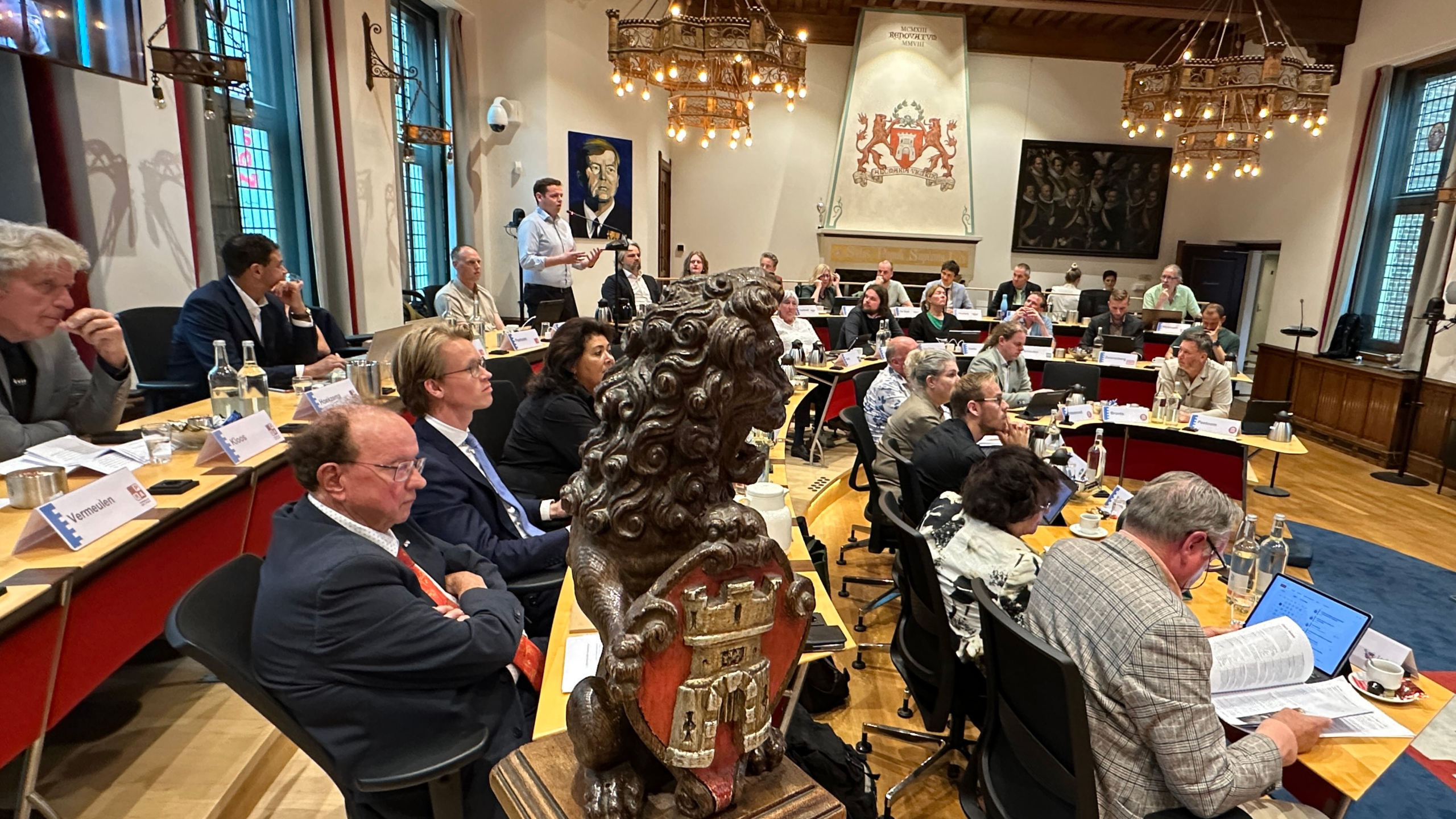 Raadsvergadering in historische zaal; man spreekt staand terwijl toehoorders zitten, met vooraan houten leeuw met wapenschild