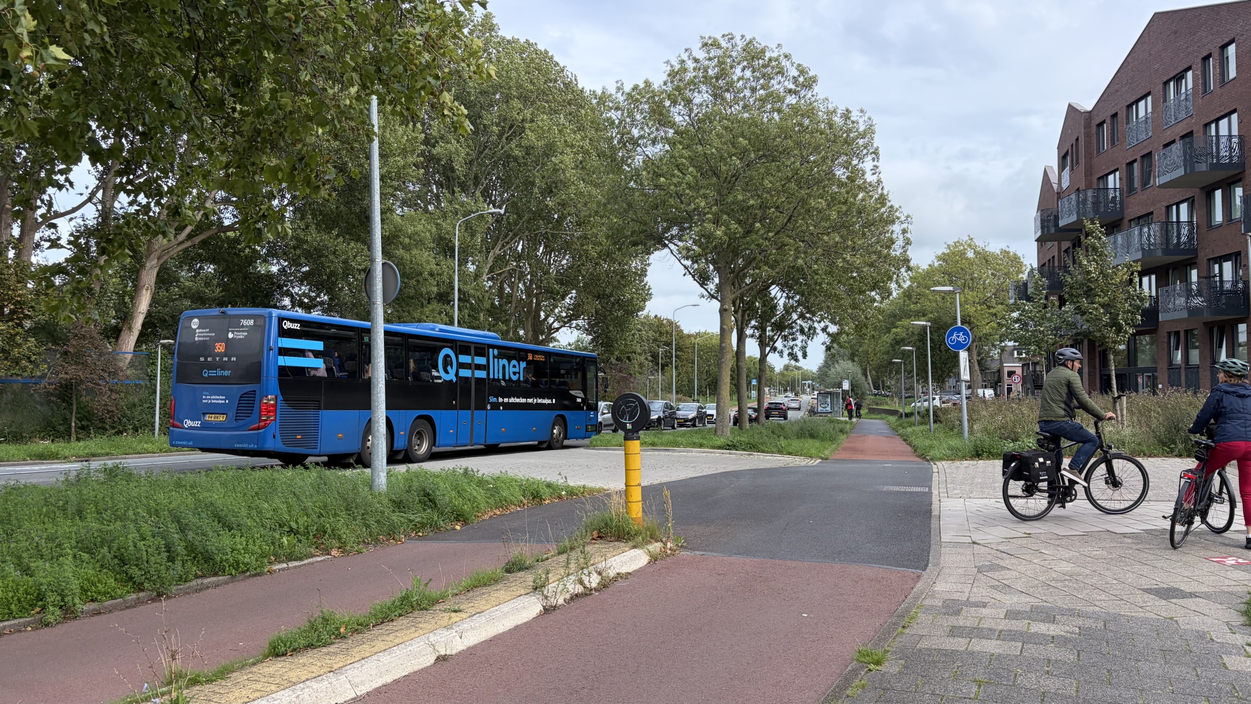 Blauwe Q-liner bus rijdt langs bomen; twee fietsers staan op het fietspad naast een modern appartementenblok