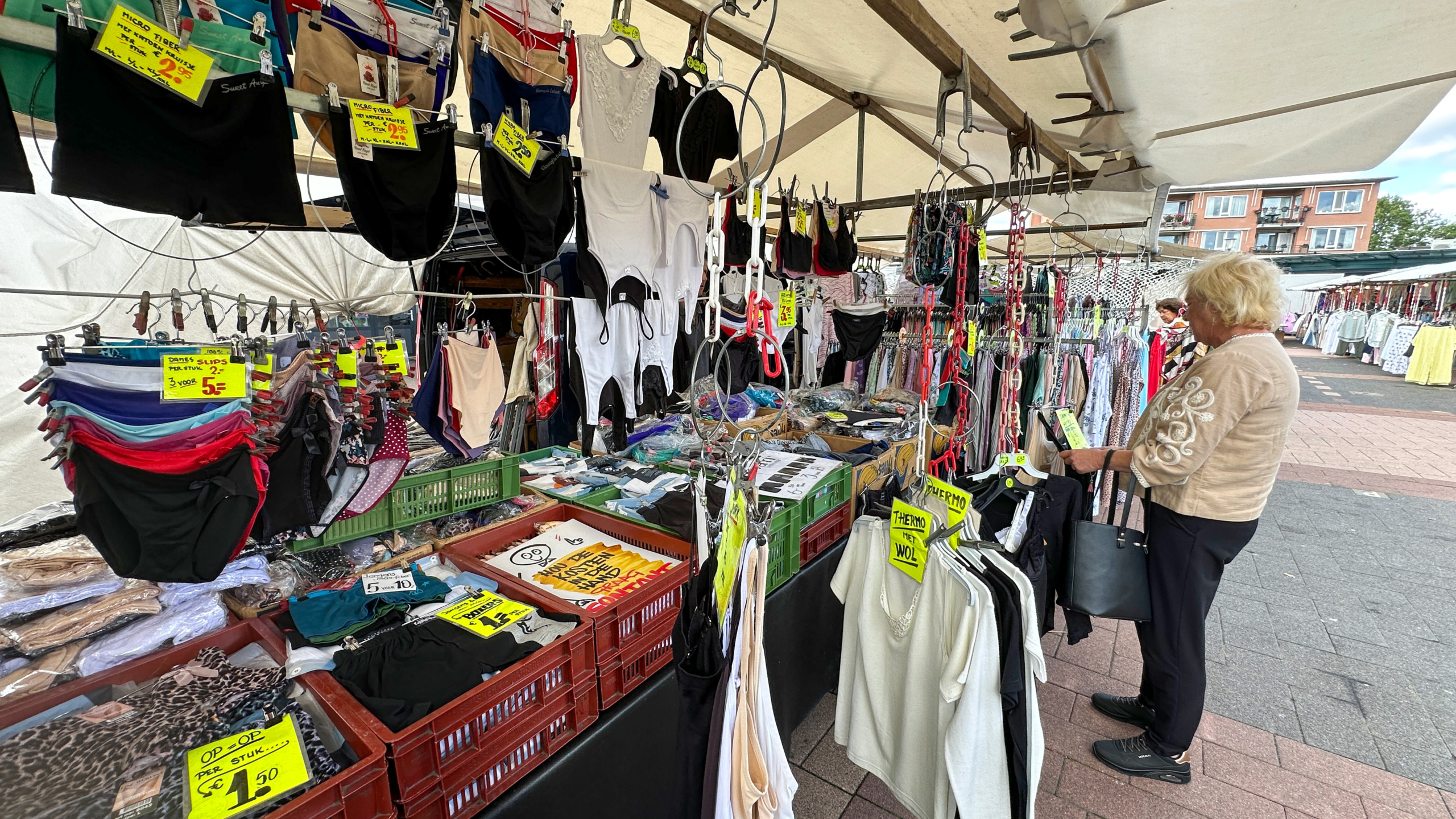 Marktkraam met ondergoed en kleding met gele prijskaartjes, terwijl een vrouw een kledingstuk bekijkt