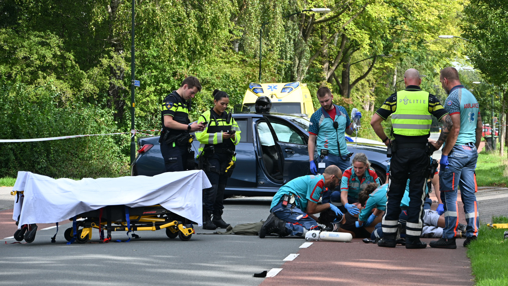 Hulpverleners en politie verlenen eerste hulp bij een verkeersongeluk naast een auto, met brancard en ambulance op de weg