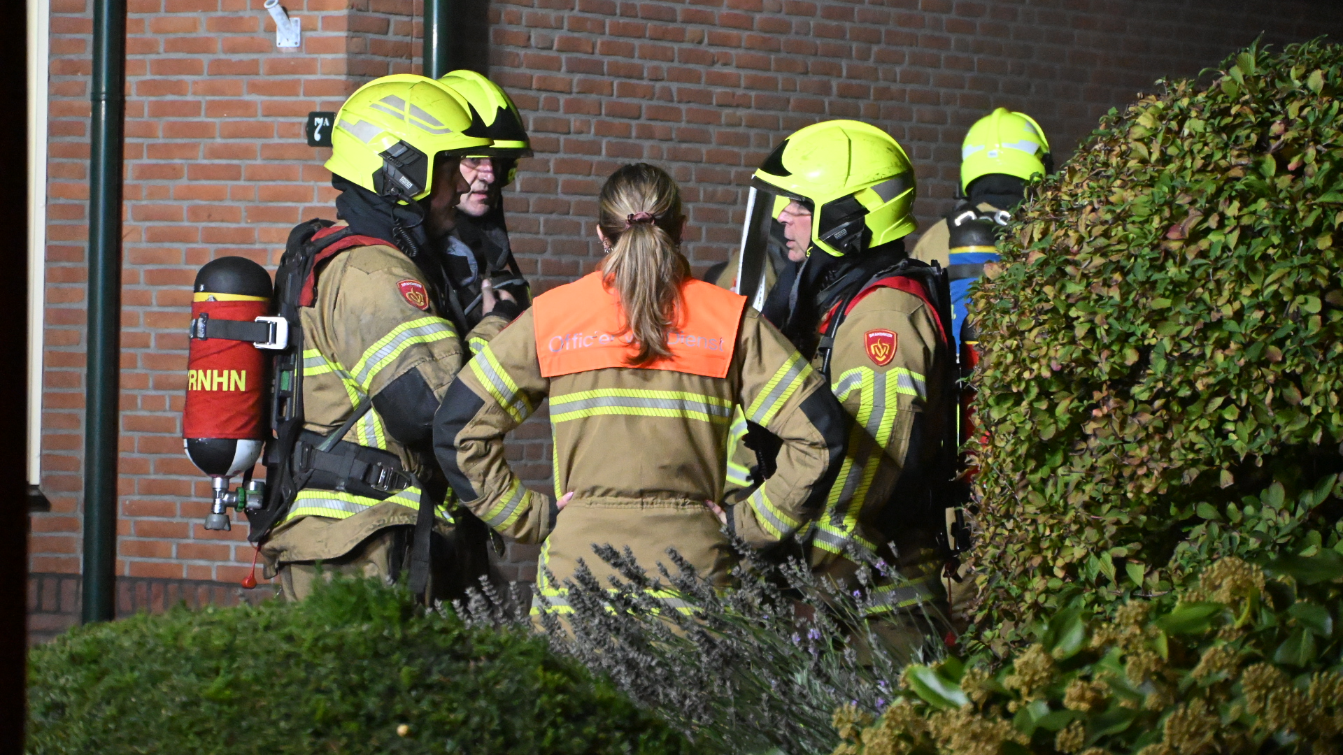 Brandweerlieden met gele helmen overleggen bij een bakstenen gevel, met ademluchtflessen en een officier van dienst