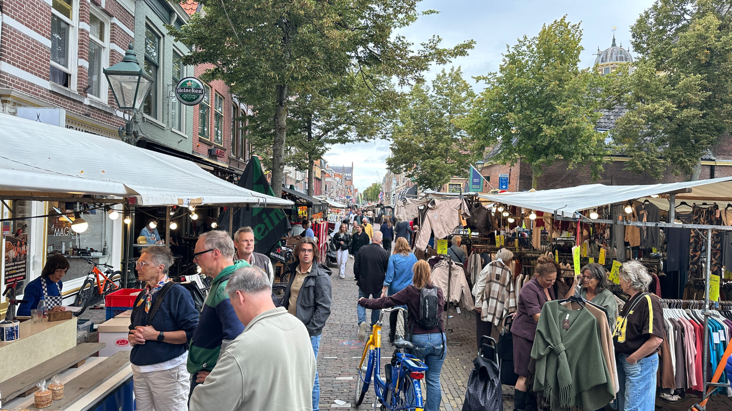 Drukke straatmarkt met kramen vol kleding en etenswaren, shoppers tussen bomen en oude gevels op een klinkerstraat