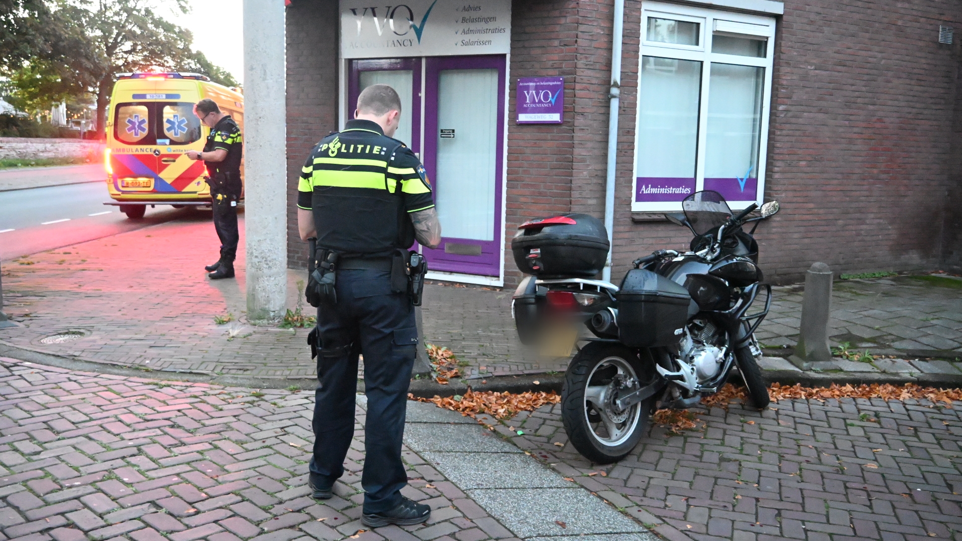 Twee agenten bij een geparkeerde motor voor een kantoor, met een ambulance met zwaailichten op de achtergrond