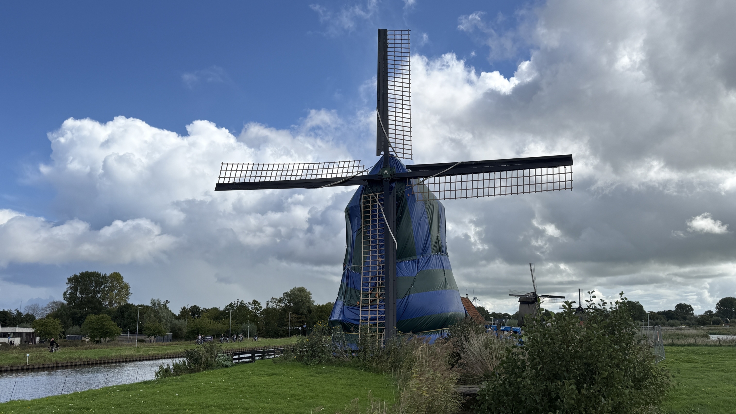 Windmolen in een groen-blauw zeilomhulsel langs een kanaal, met wieken tegen een wolkenlucht