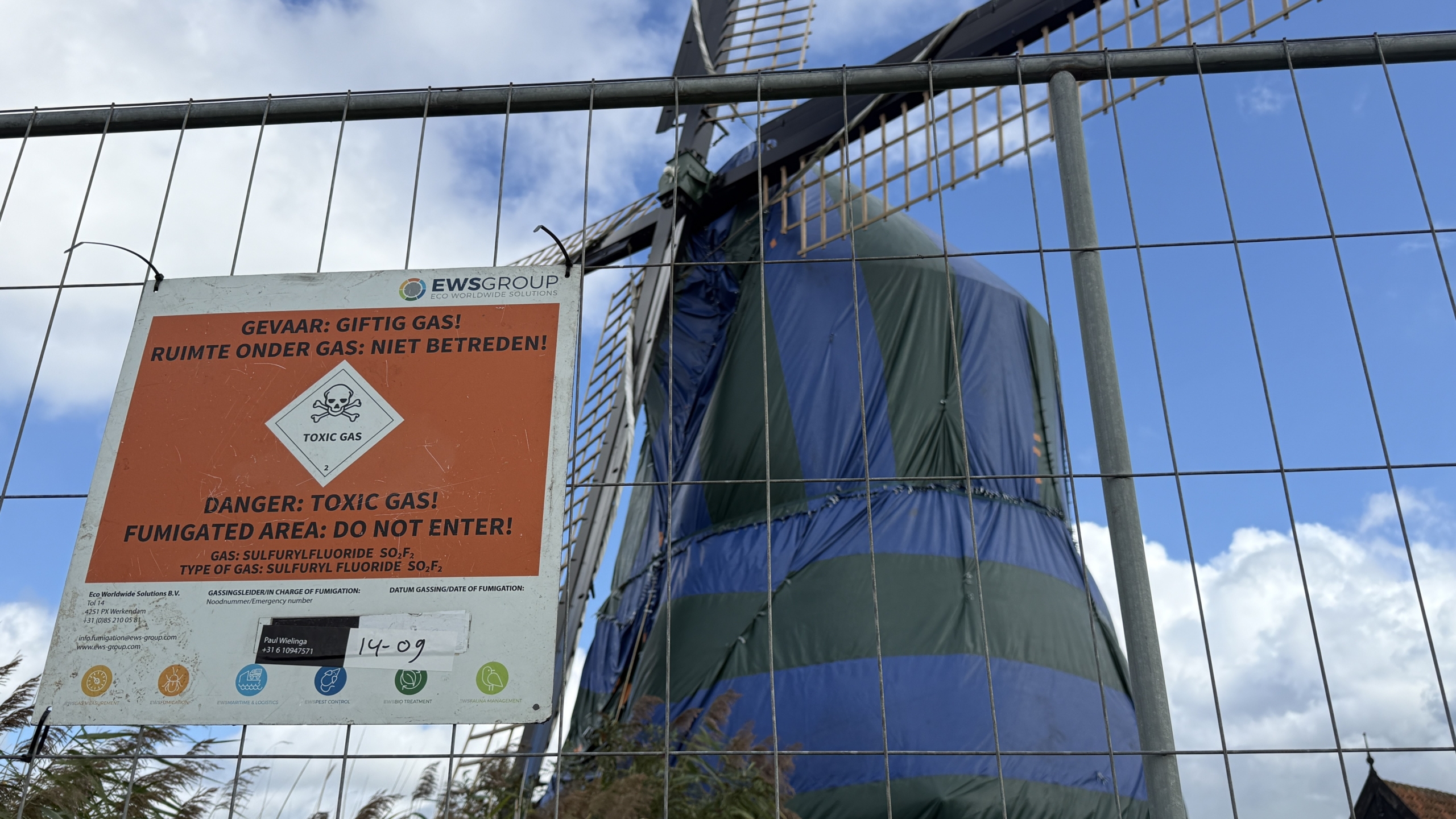 Waarschuwingsbord “Gevaar: giftig gas, niet betreden” aan hek, met afgedekte molen op de achtergrond