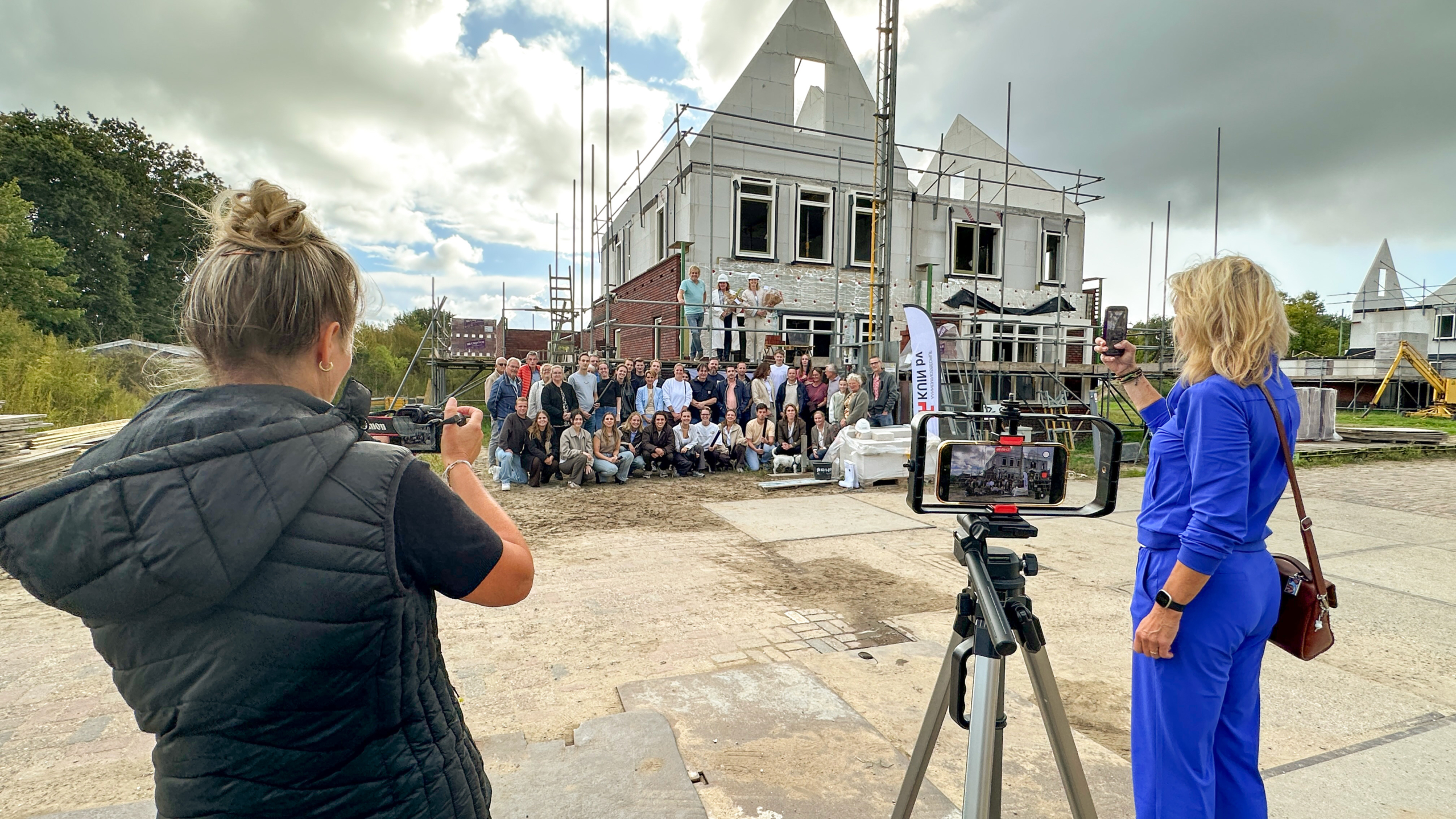 Groepsfoto bij een huis in aanbouw met steigers; twee vrouwen maken foto’s, één met smartphone op statief