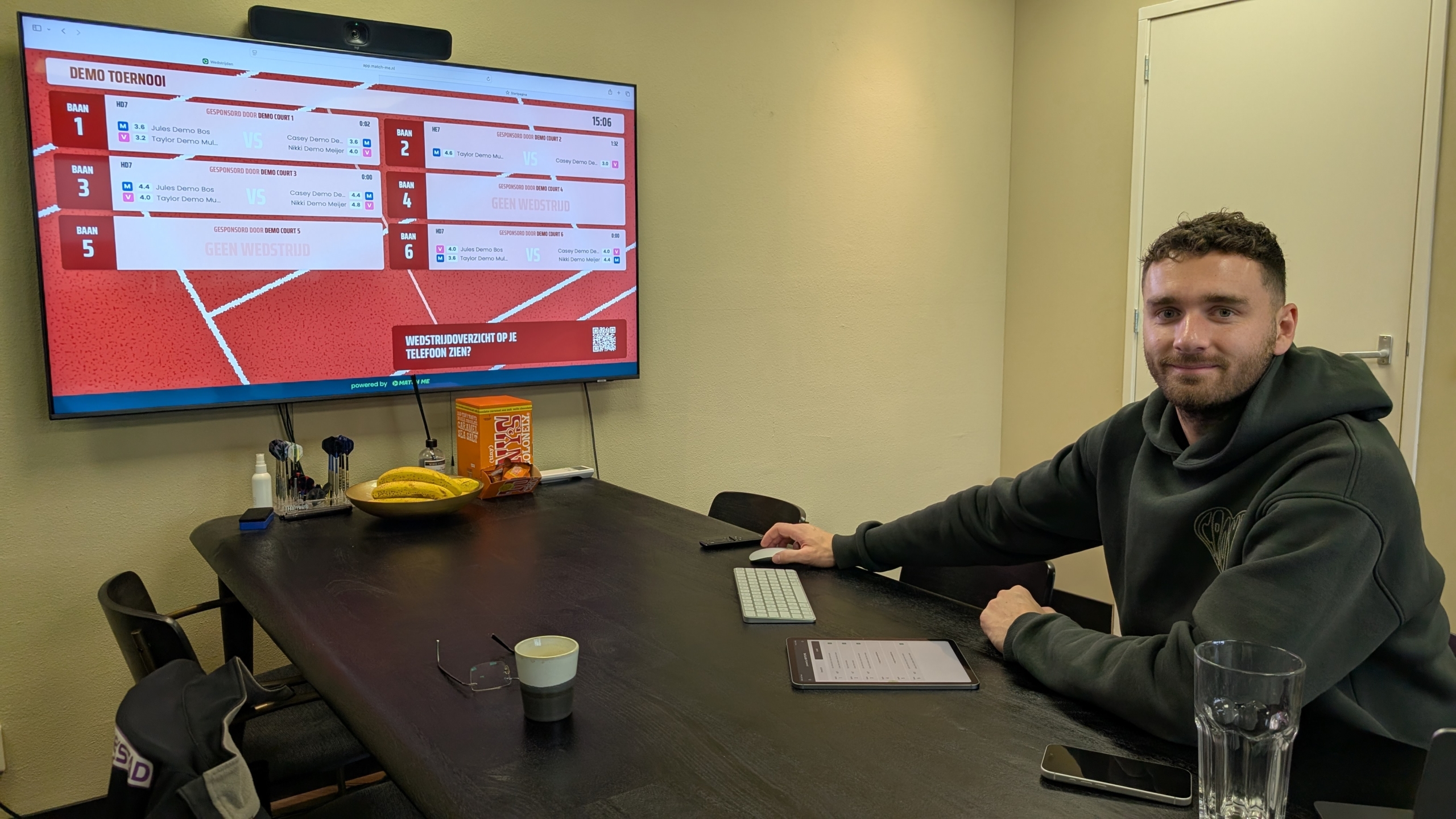 Man zit aan tafel met toetsenbord en tablet; op groot scherm staat een demo-toernooioverzicht met banen en wedstrijden