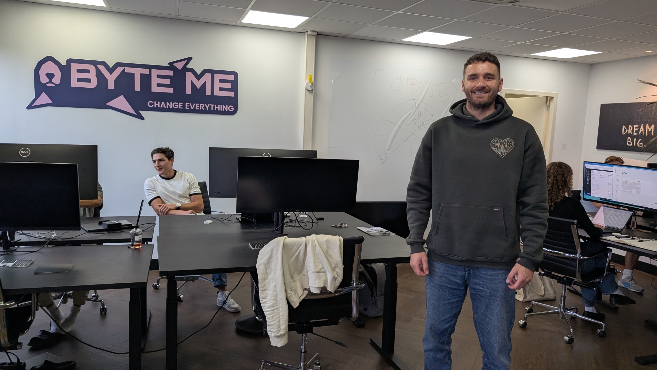 Man in hoodie staat in een kantoor met werkplekken; op de muur hangt het logo Byte Me met de tekst Change Everything