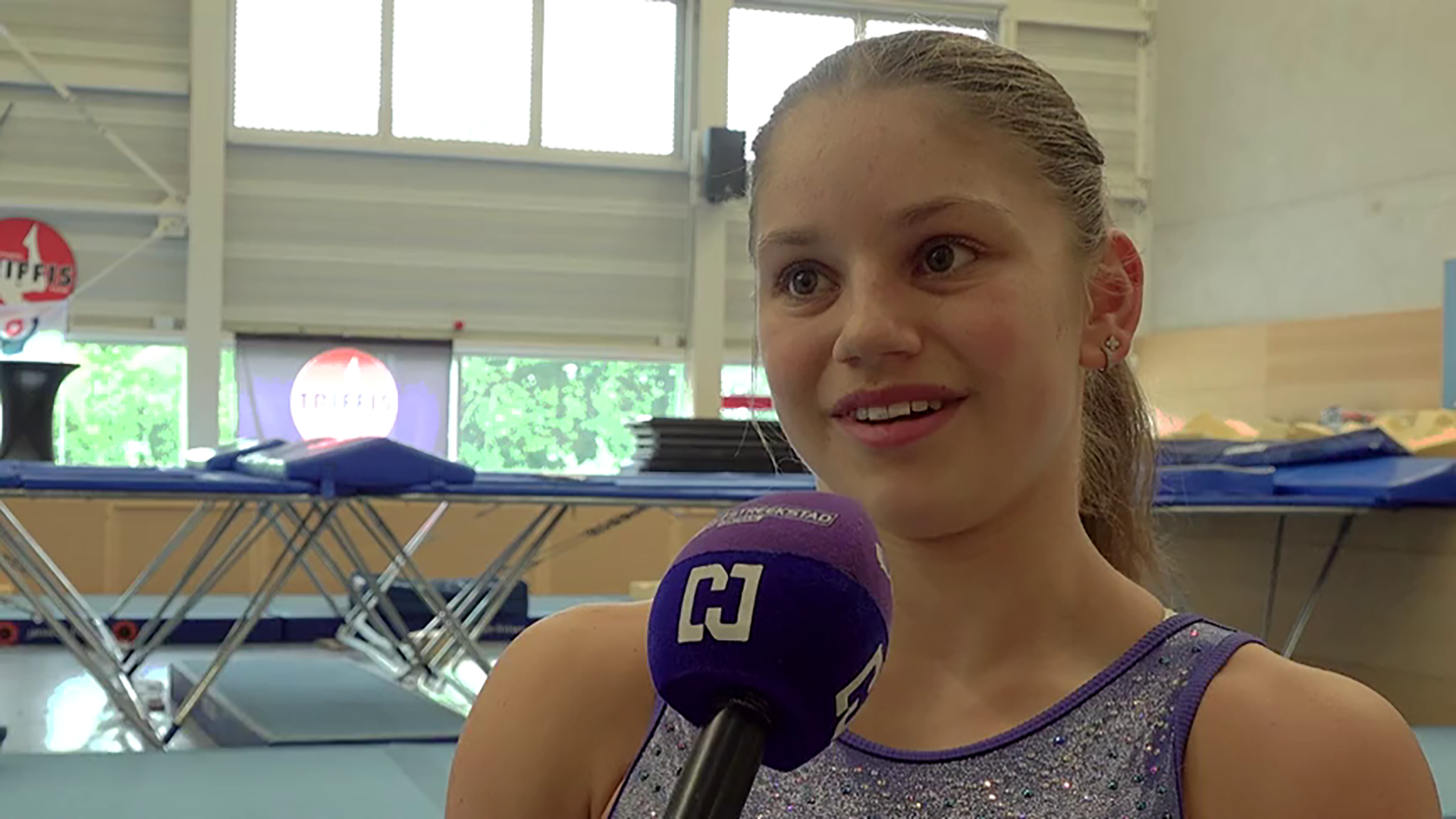 Turnster in sporthal geeft interview in close-up, met microfoon op de voorgrond en trampolines op de achtergrond