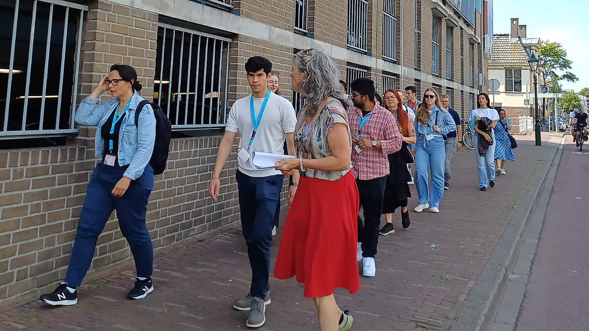 Groep mensen met blauwe naambadges loopt langs een bakstenen gebouw, geleid door een vrouw met grijs haar en rode rok