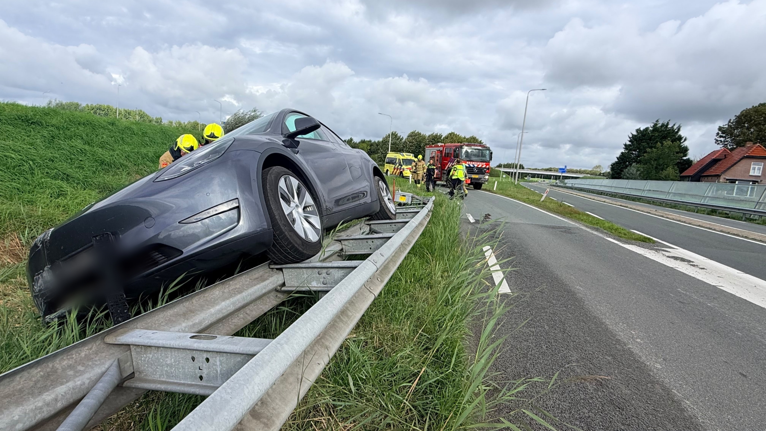 Grijze auto staat scheef op de vangrail langs een afrit, met brandweer en ambulance op de achtergrond