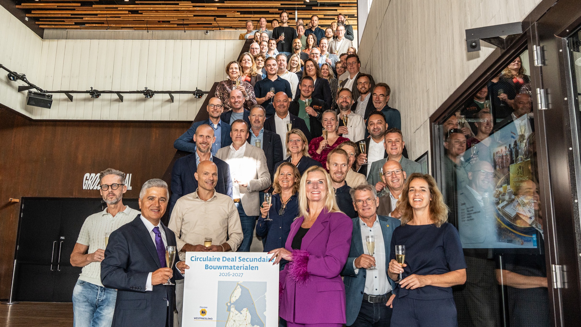 Groepsfoto van tientallen mensen op een trap met champagneglazen, bij een bord “Circulaire Deal Secundaire Bouwmaterialen”