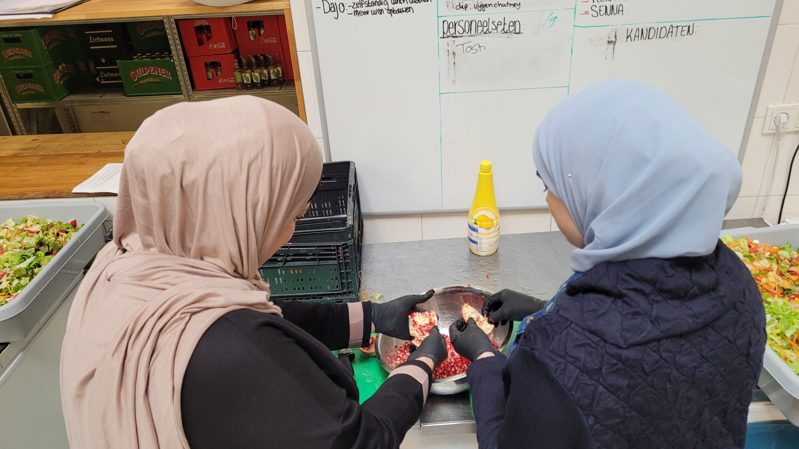 Twee vrouwen met hoofddoek maken granaatappel schoon boven een kom in een keuken, met bakken salade op de achtergrond