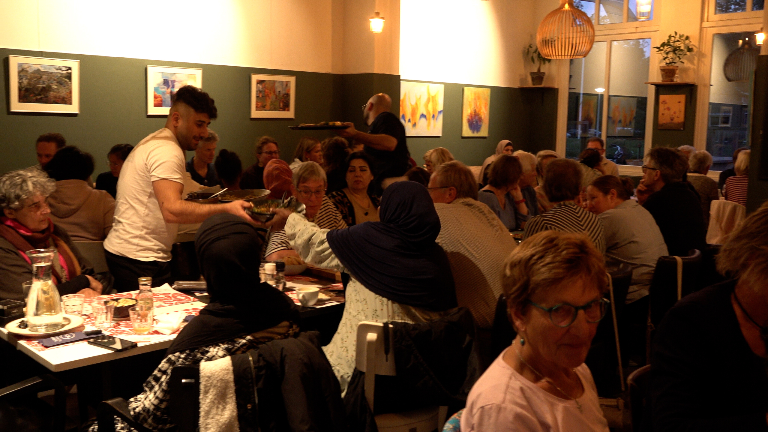 Drukke eetzaal met veel gasten aan lange tafels terwijl een serveerder schalen eten uitdeelt