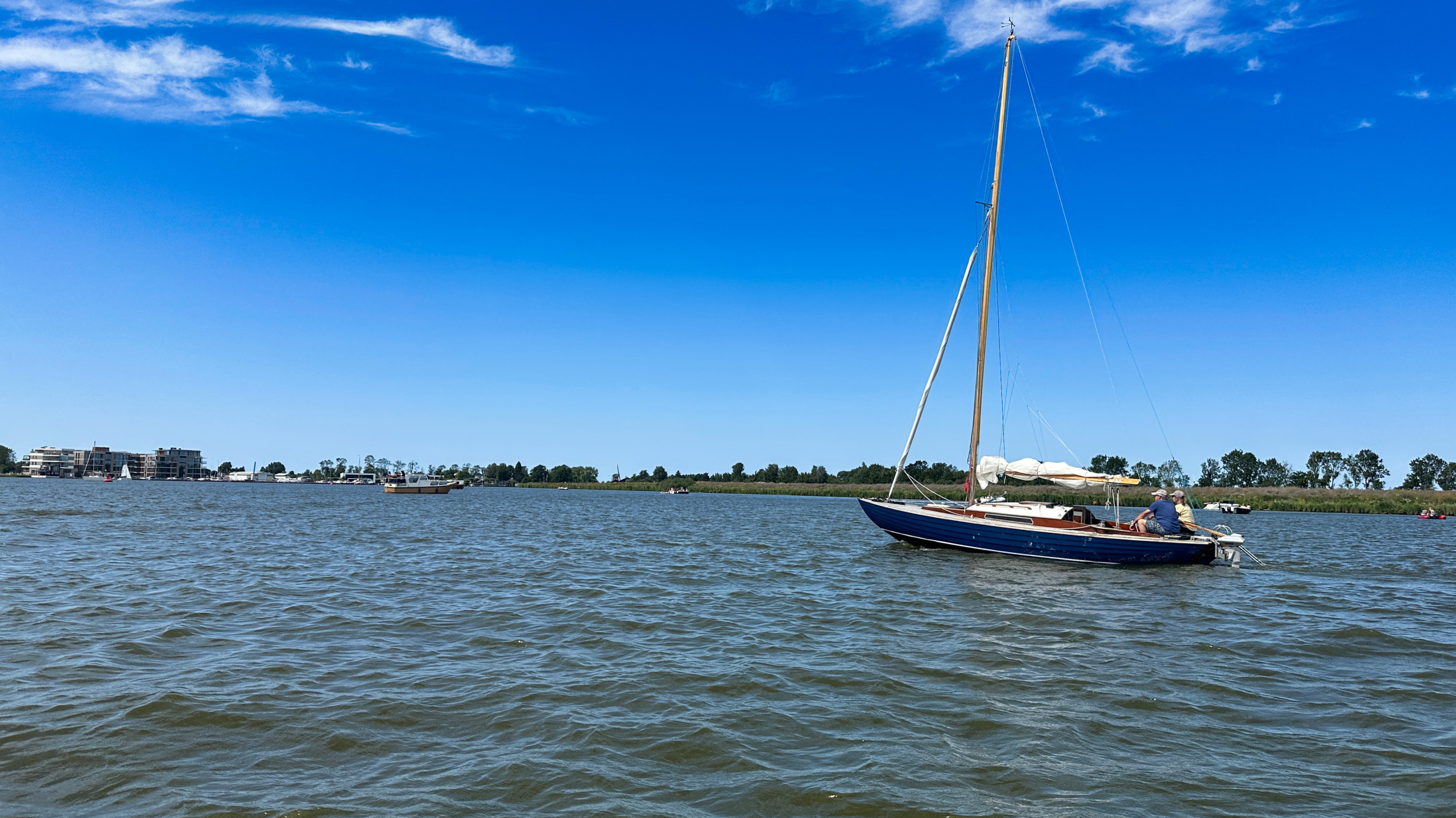Blauwe zeilboot met gestreken zeil op een meer, met twee mensen aan boord en een heldere blauwe lucht