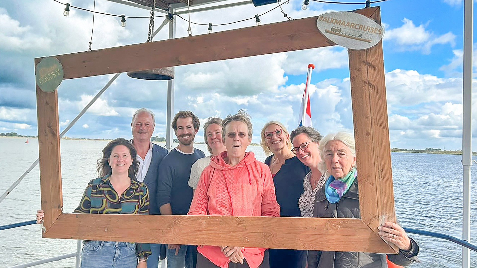 Groep mensen op een boot poseert met een groot houten fotolijstkader; op het bord staat “#ALKMAARCRUISES”