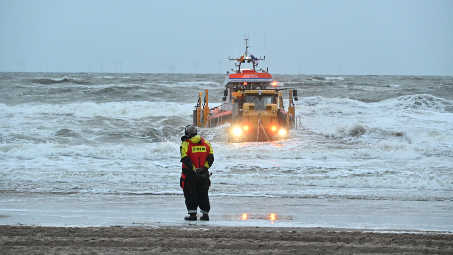Reddingsboot vaart door ruwe zee naar strand terwijl een KNRM-hulpverlener toekijkt