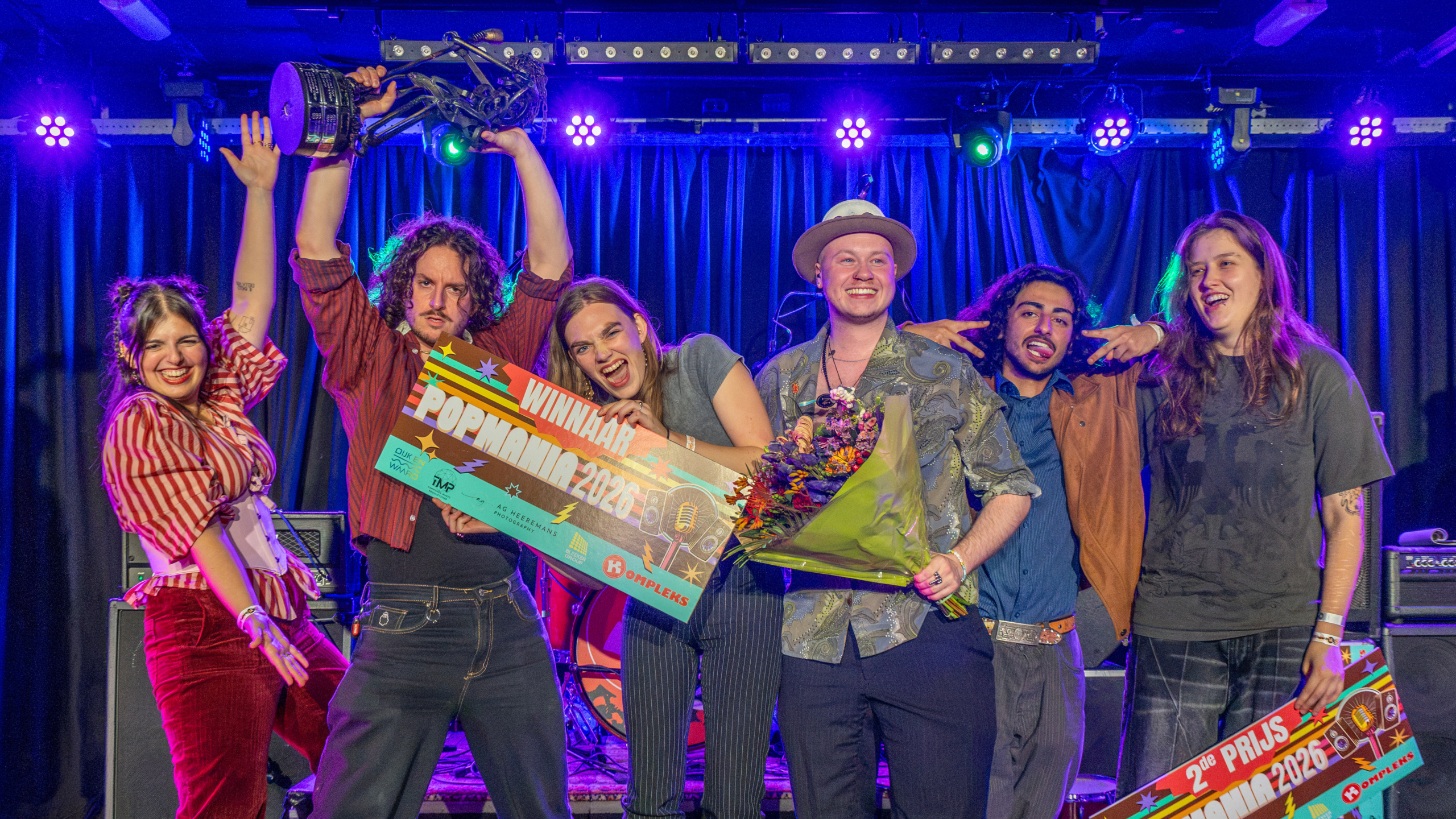 Groep muzikanten viert op het podium met trofee, bloemen en bord met tekst WINNAAR Popmania 2026