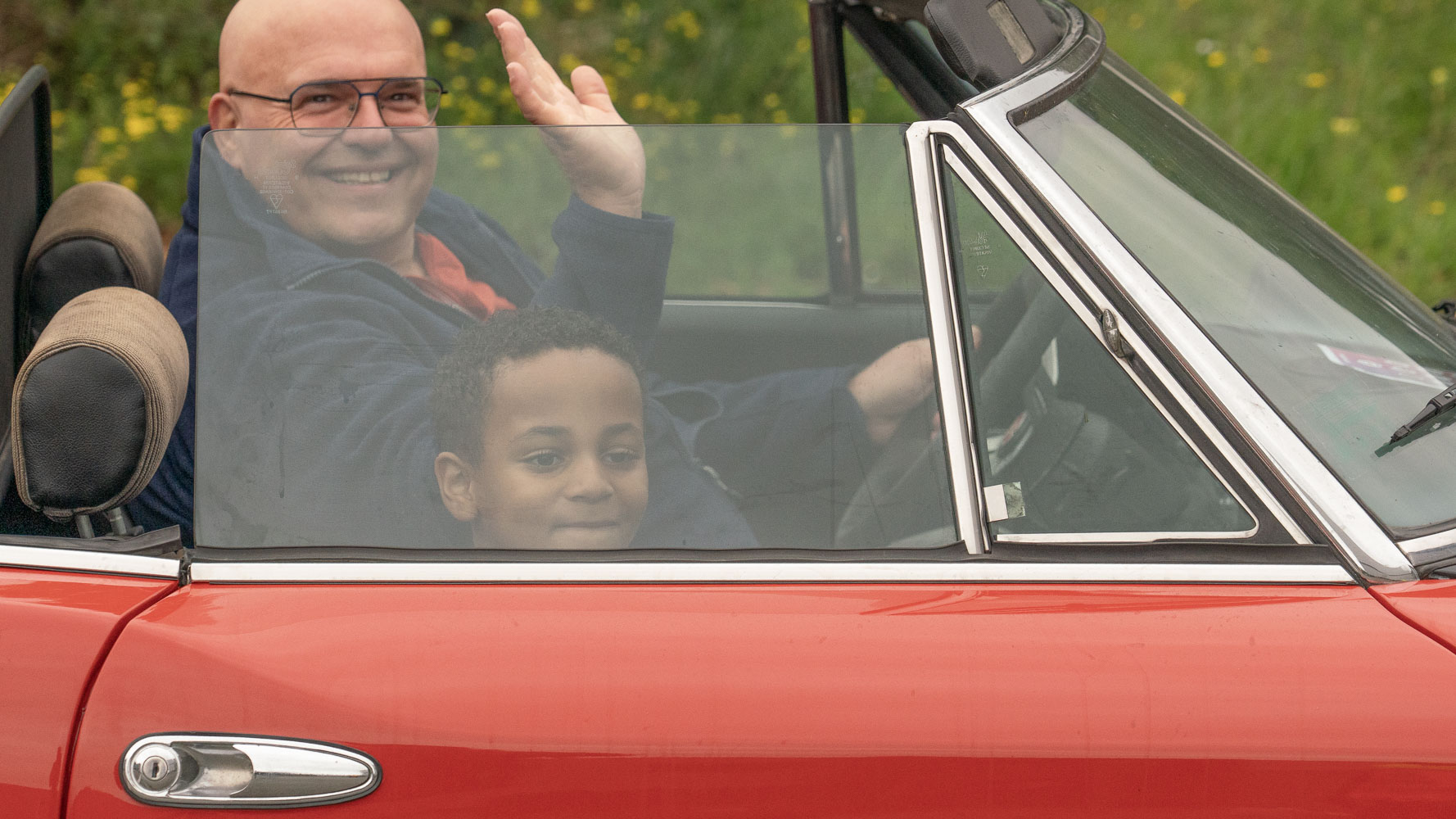 Man zwaait vanuit een rode cabriolet, met een kind op de passagiersstoel, tegen een groene achtergrond