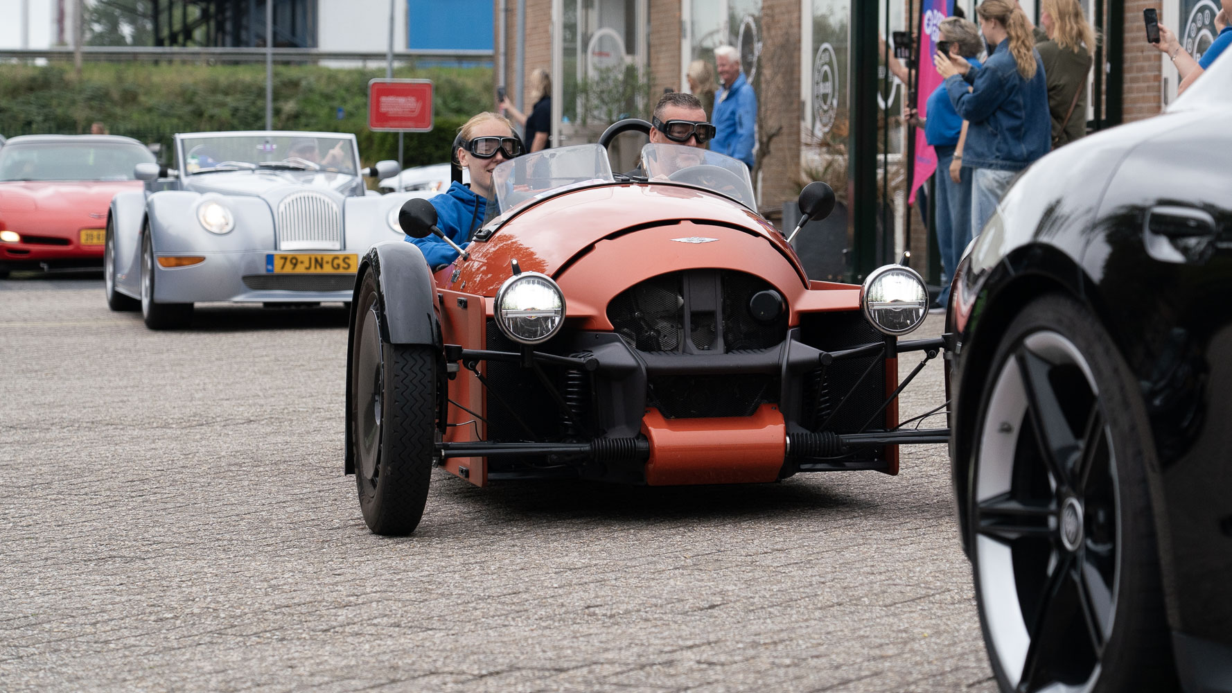 Oranje open sportauto met twee inzittenden met goggles rijdt in stoet langs toeschouwers, met andere cabrio’s erachter