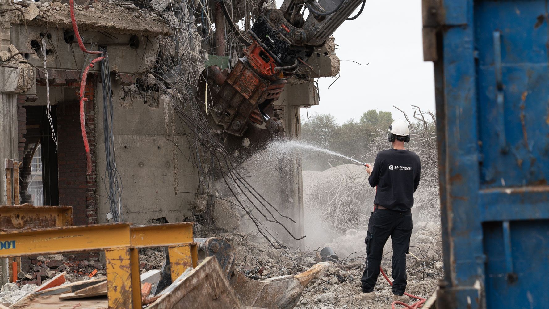 Bouwvakker in helm besproeit puin en stof terwijl een sloopkraan een betonnen gebouw afbreekt
