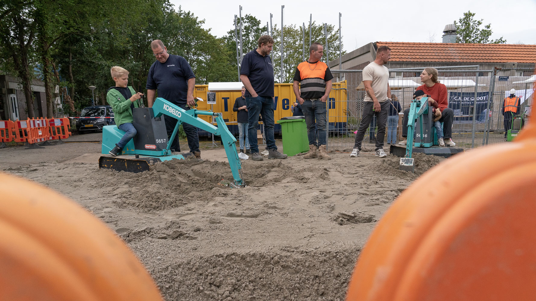 Kind en vrouw bedienen minigravers in een zandbak, terwijl volwassenen toekijken op een afgezette bouwplaats