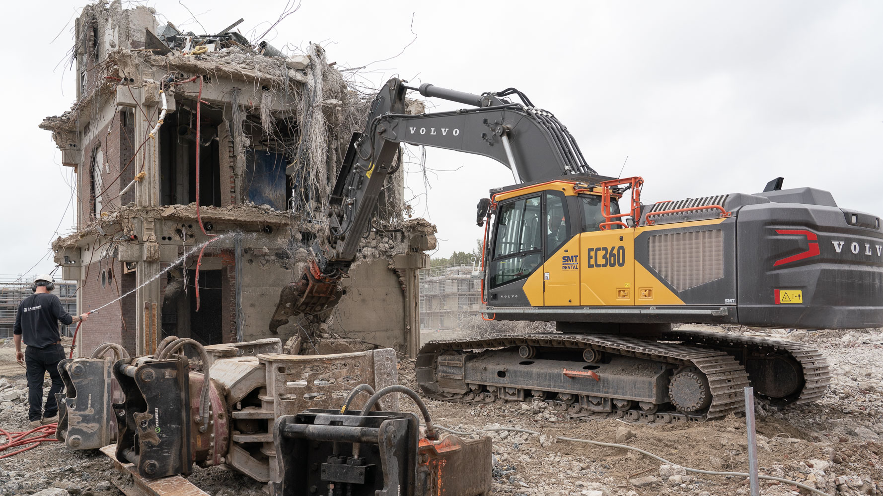 Graafmachine sloopt een gebouw; beton en kabels hangen los, met puin op de grond en een arbeider links