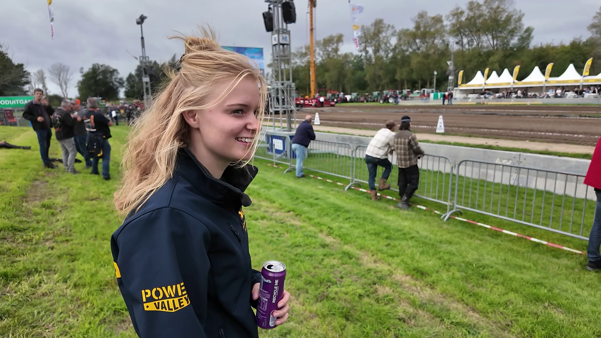 Lachend blond meisje met blikje drinken bij een modderige racebaan, met toeschouwers en tenten op de achtergrond
