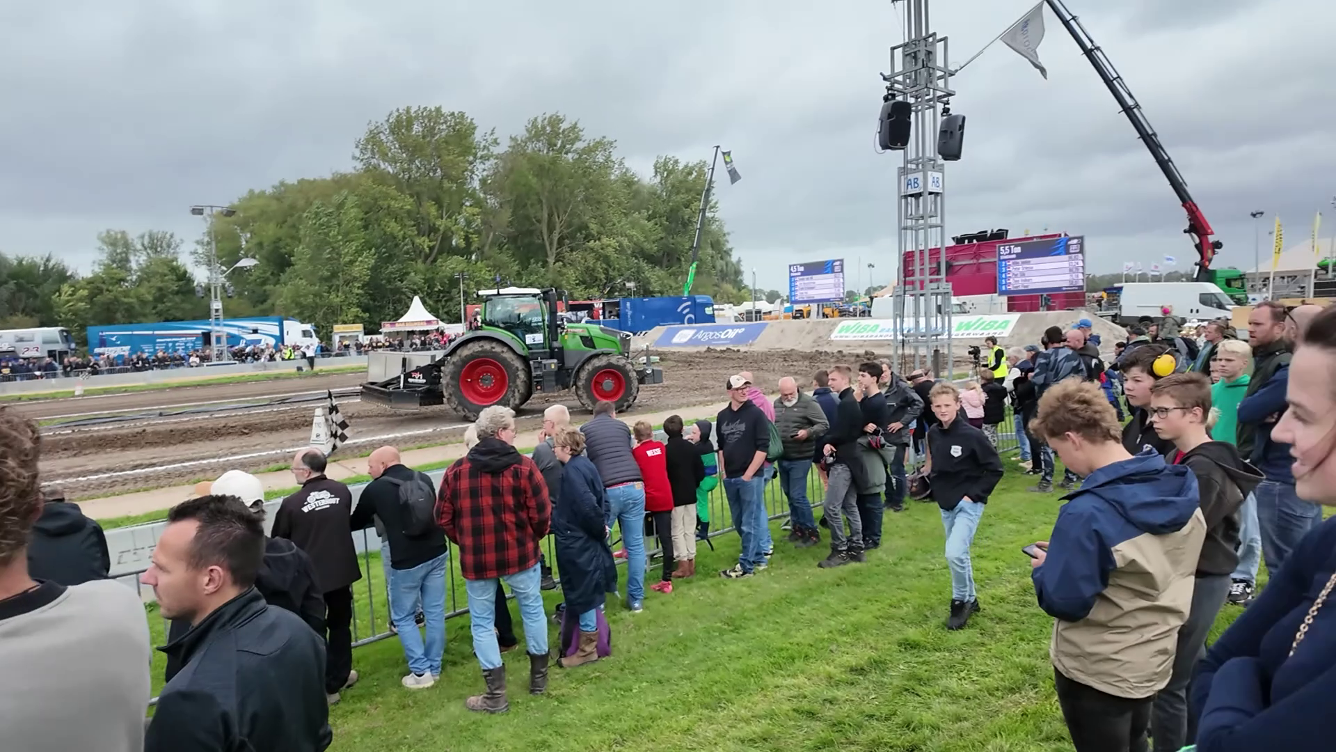 Toeschouwers langs een modderige trekkerbaan terwijl een groene tractor voorbijrijdt bij een buitenevenement