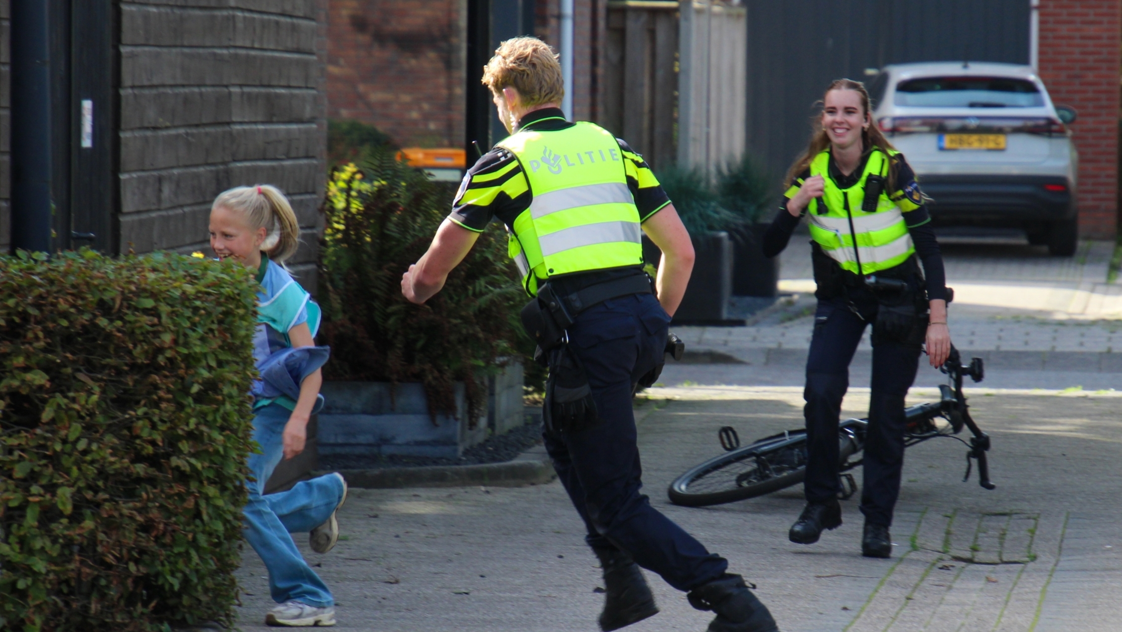 Twee politieagenten in gele hesjes rennen lachend achter een meisje aan in een woonstraat, met een fiets op de grond