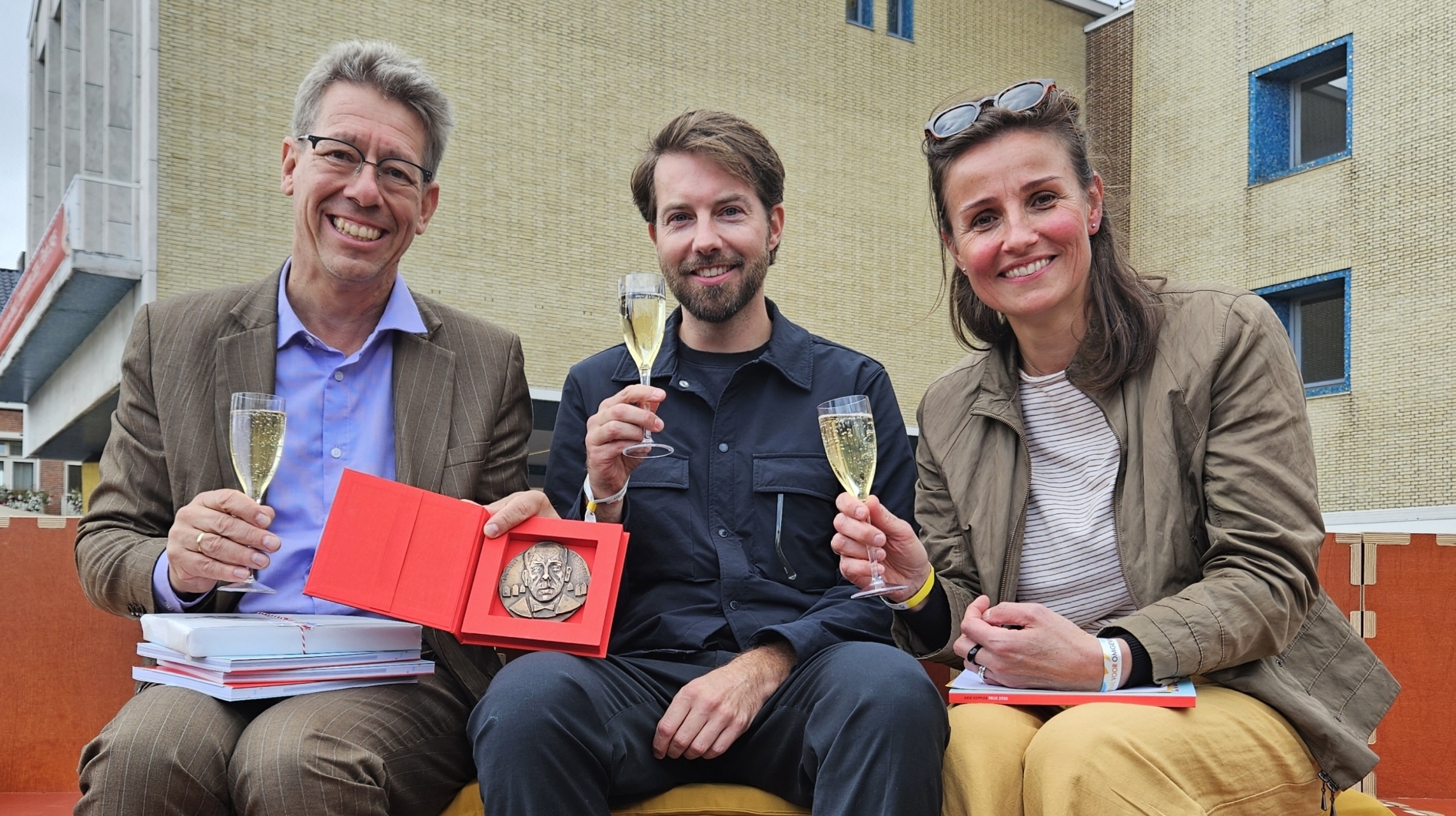 Drie mensen proosten met champagne; de man links toont een medaille in rood doosje, zittend op een buitenbank