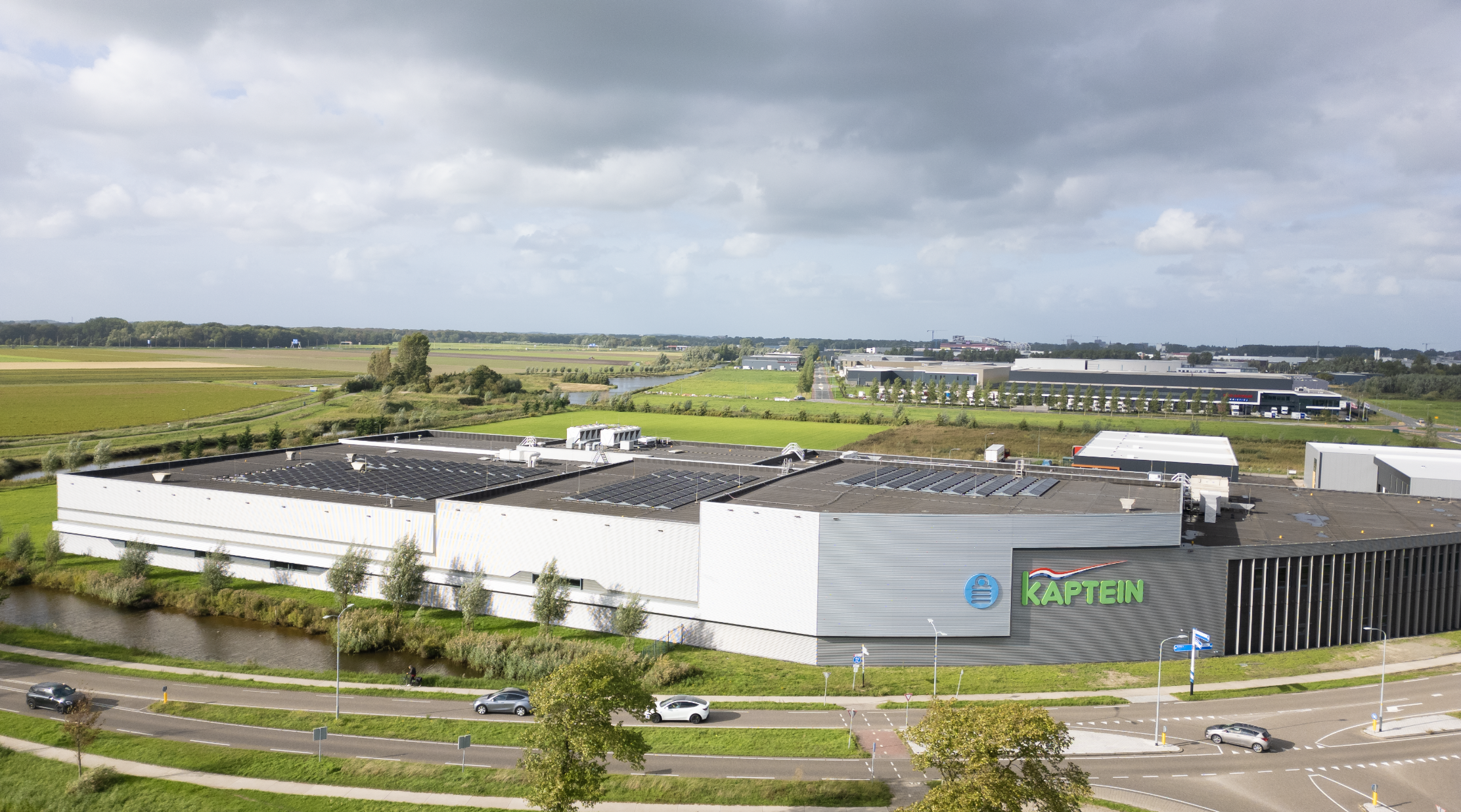 Luchtfoto van een fabriekspand met zonnepanelen en het logo Kaptein, naast een kanaal en weg, met weilanden eromheen