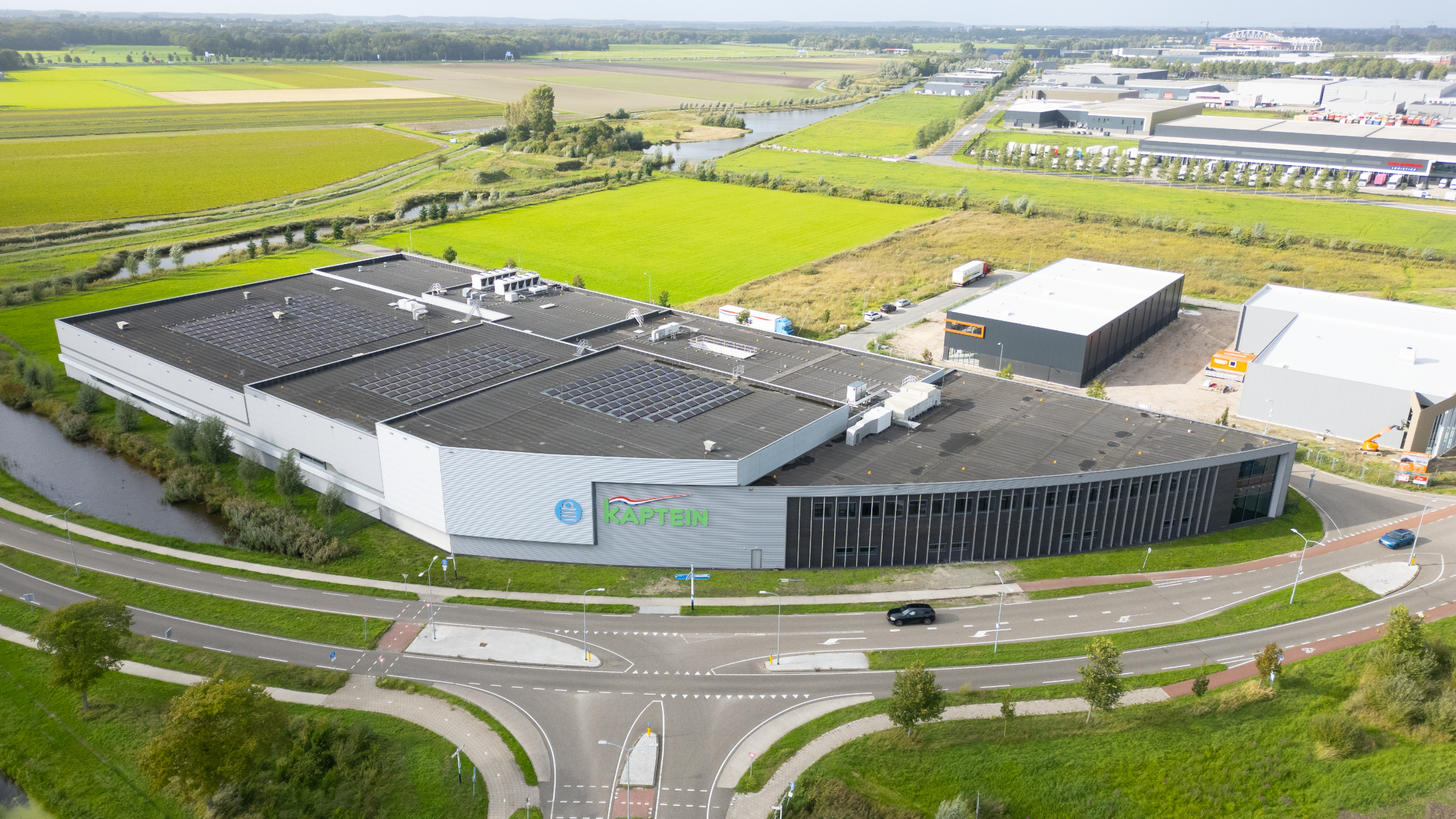 Luchtfoto van bedrijfsgebouw Kaptein met zonnepanelen op het dak, langs een kruispunt tussen groene velden