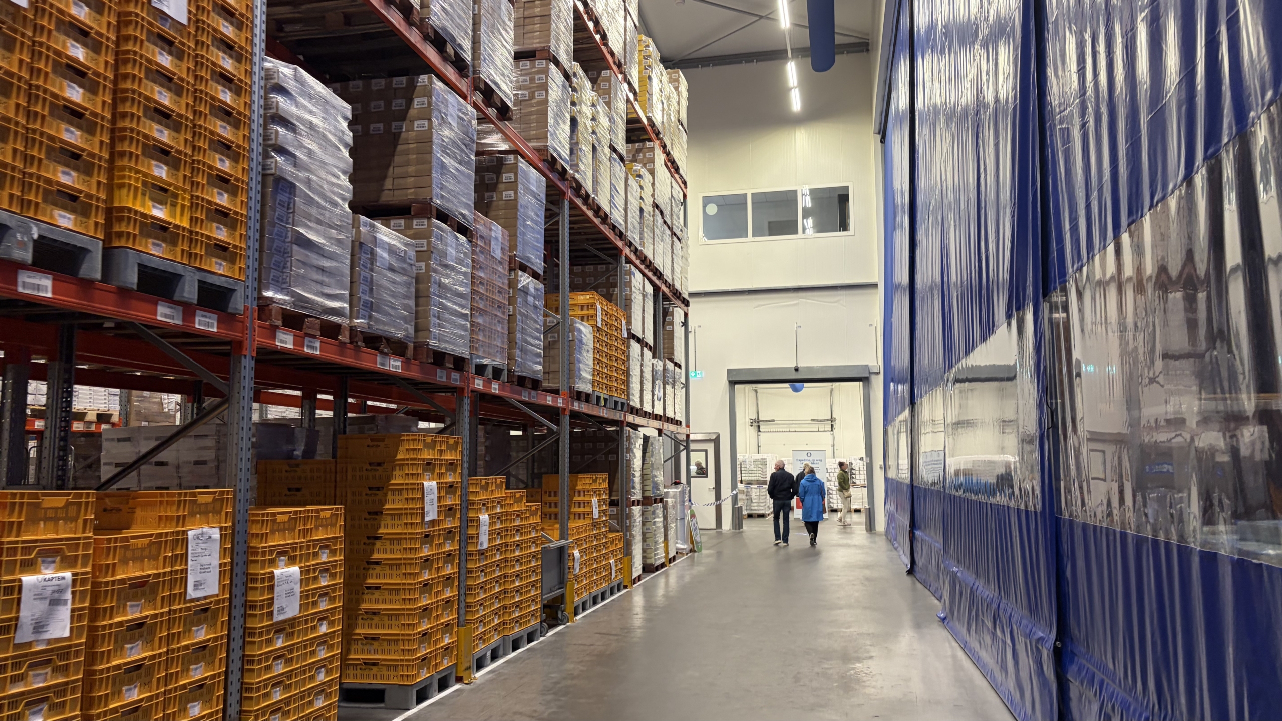 Gangpad in magazijn met hoge stellingen vol pallets en gele kratten, met mensen die richting een deur lopen