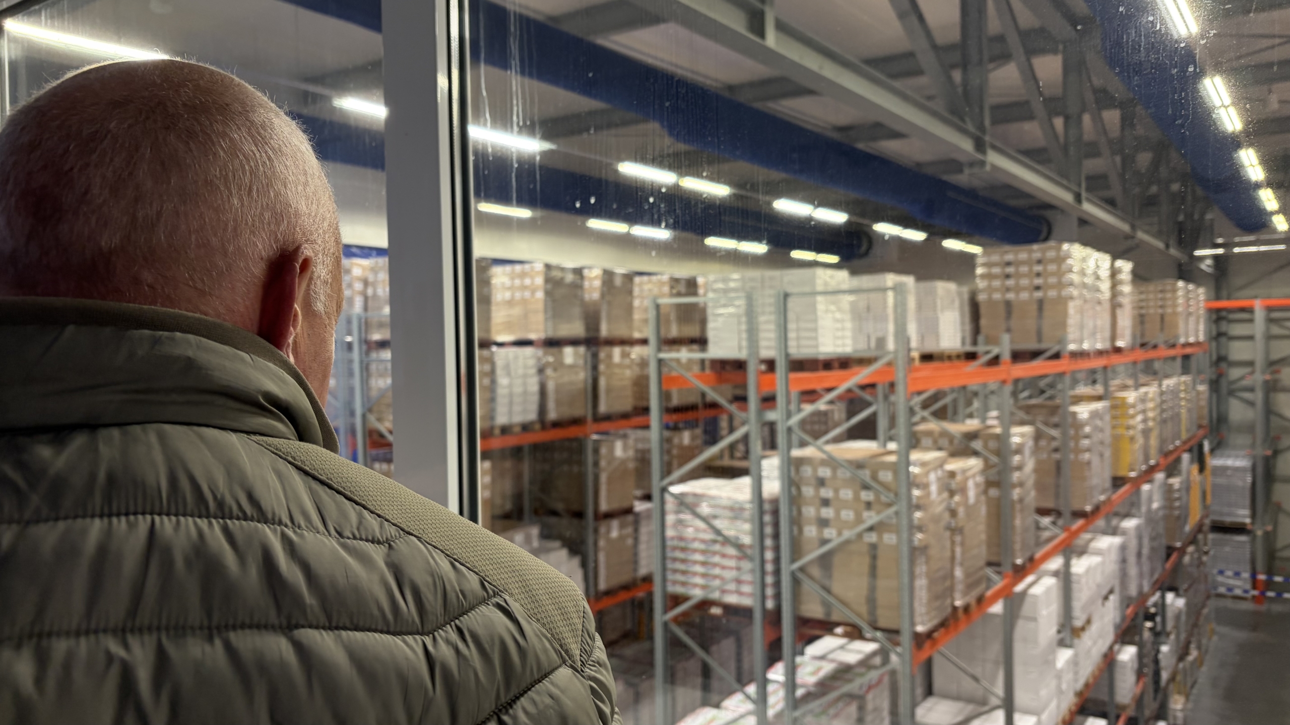 Man met kort haar kijkt door een raam uit over magazijnstellingen vol pallets en dozen in een distributiecentrum