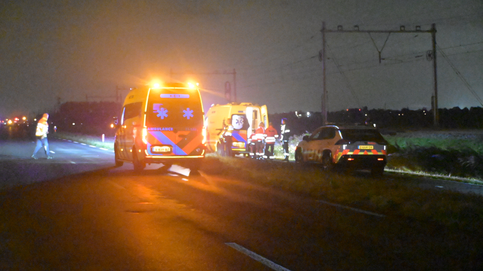 Ambulance met zwaailichten en hulpverleners langs een donkere weg, met een auto in de berm